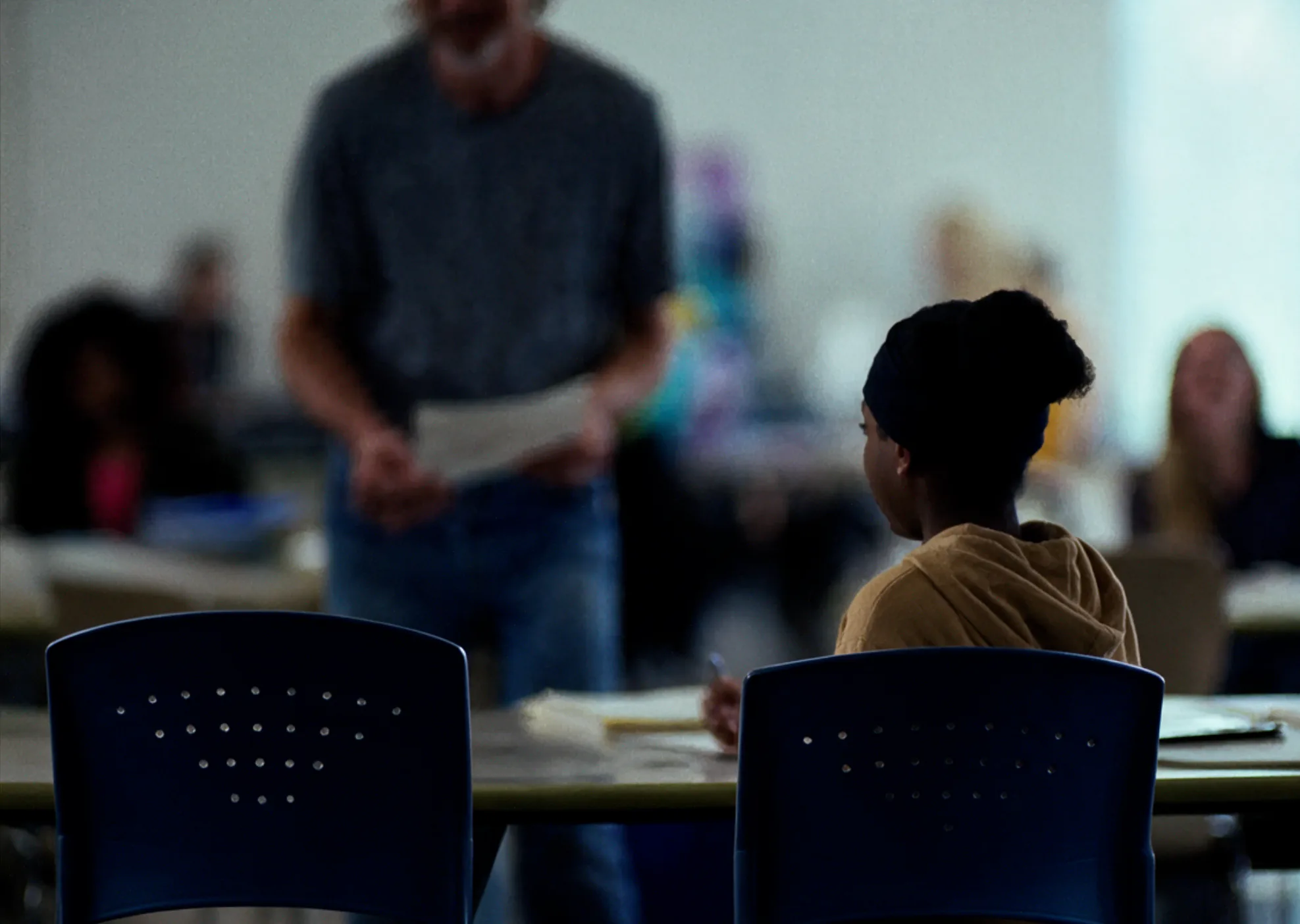 Two empty blue chairs at a classroom table in the foreground, one beside a student in a mustard hoodie writing while a teacher stands holding papers, out of focus behind them