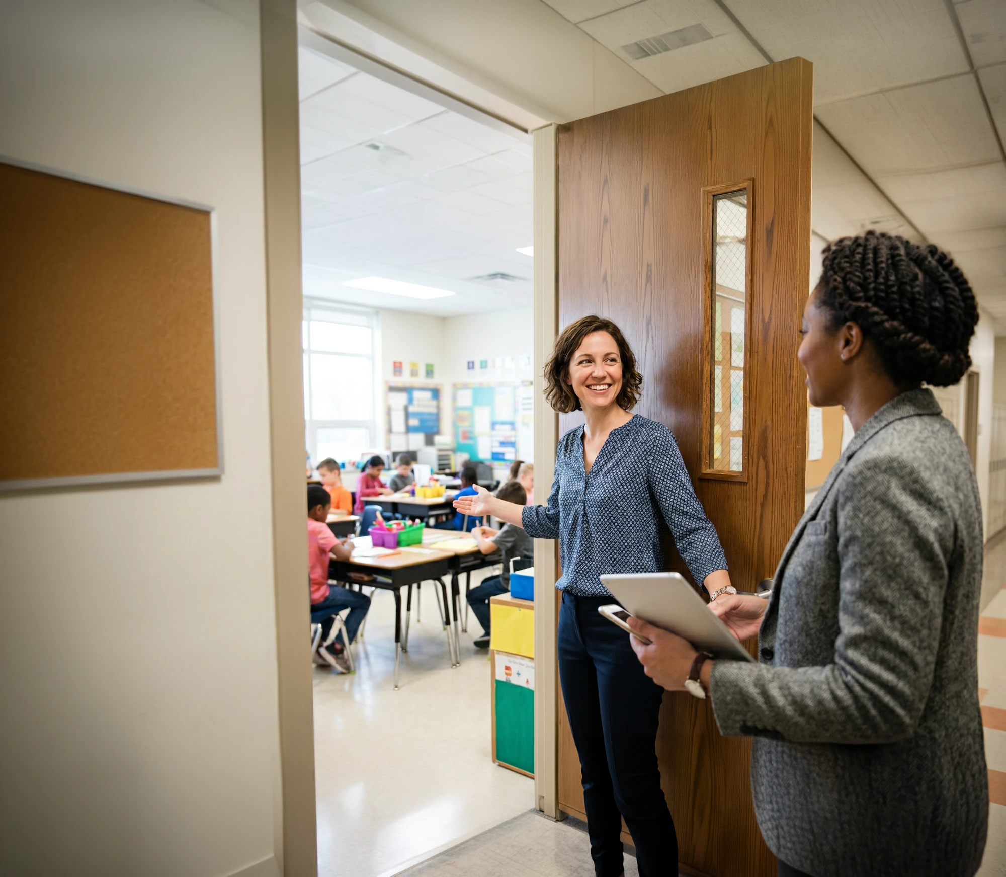 A smiling teacher holds her classroom door open and waves in a colleague in a gray blazer carrying a tablet and phone, students working at tables in the bright room behind her