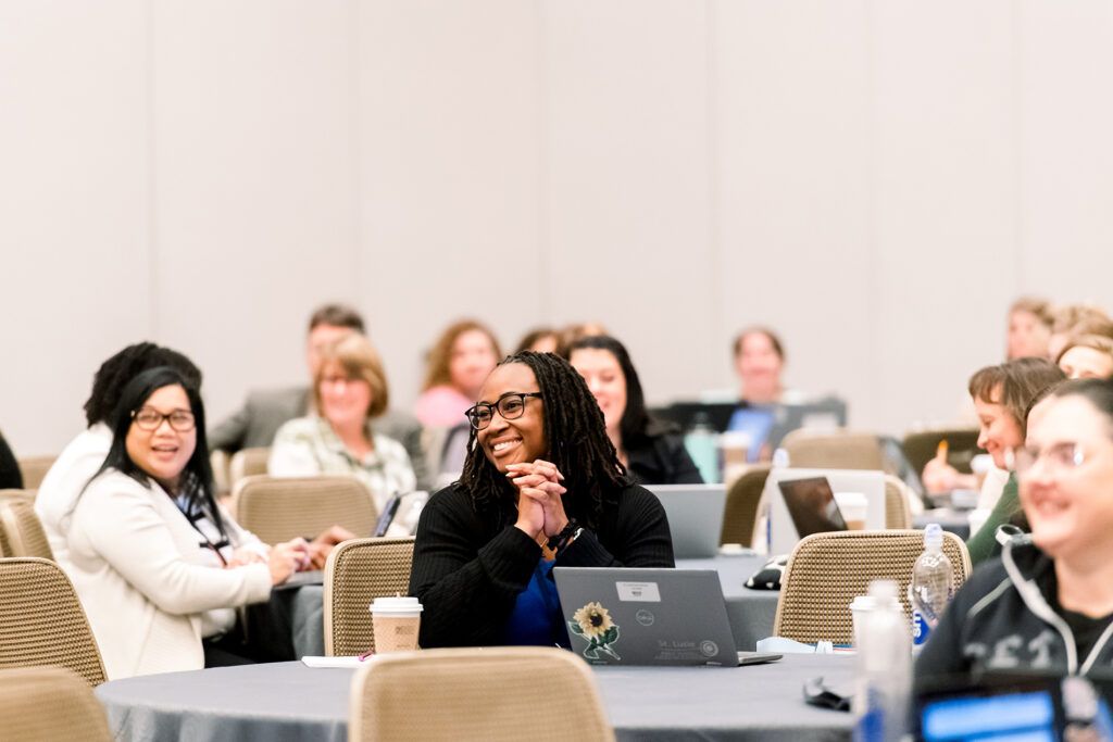An educator in glasses, her hands clasped and smiling, sits among attendees at round tables with laptops during a conference session