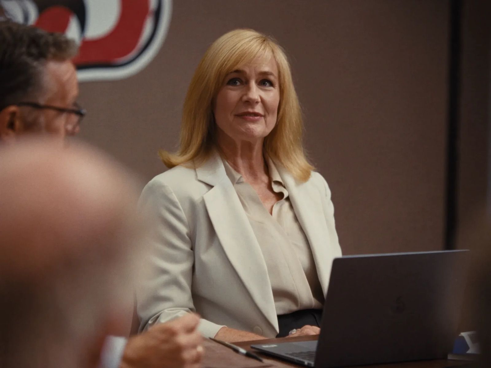 A district leader with shoulder-length blonde hair and a cream blazer sits at a conference table behind a laptop, smiling as a colleague speaks, a district logo on the wall behind