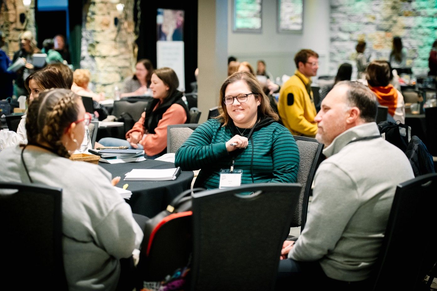 At a round table in a busy workshop hall, a smiling educator in glasses and a striped green sweater, pen in hand, talks with two colleagues, other tables of educators out of focus behind them