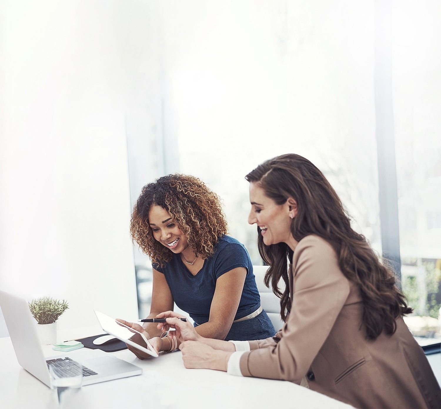 Two smiling educators at a white desk in a bright office, one in a navy top and one in a tan blazer, look over a tablet together beside an open laptop