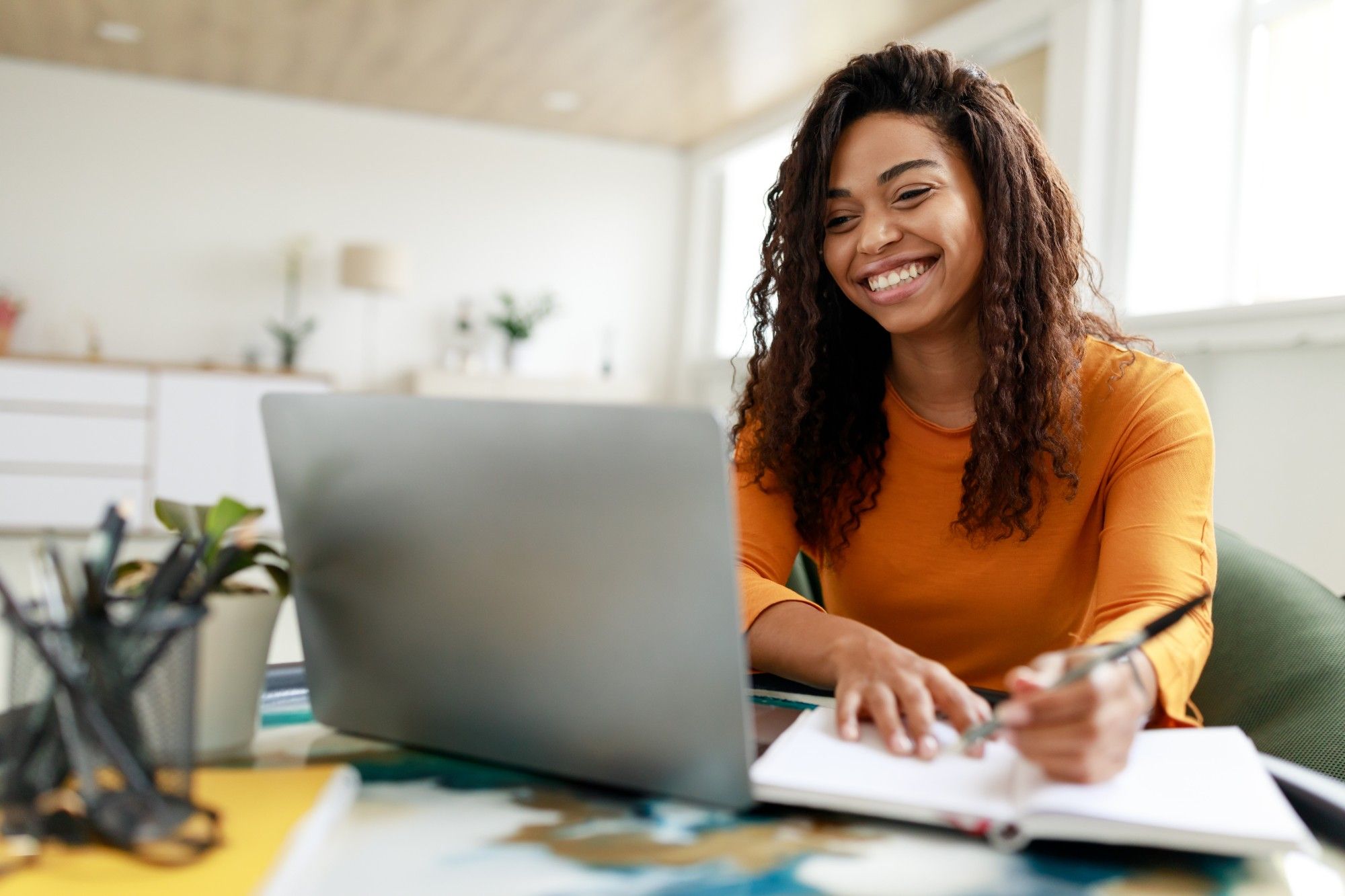 A smiling educator with long curly hair, in an orange top, takes notes in a notebook while working at a laptop in a bright room