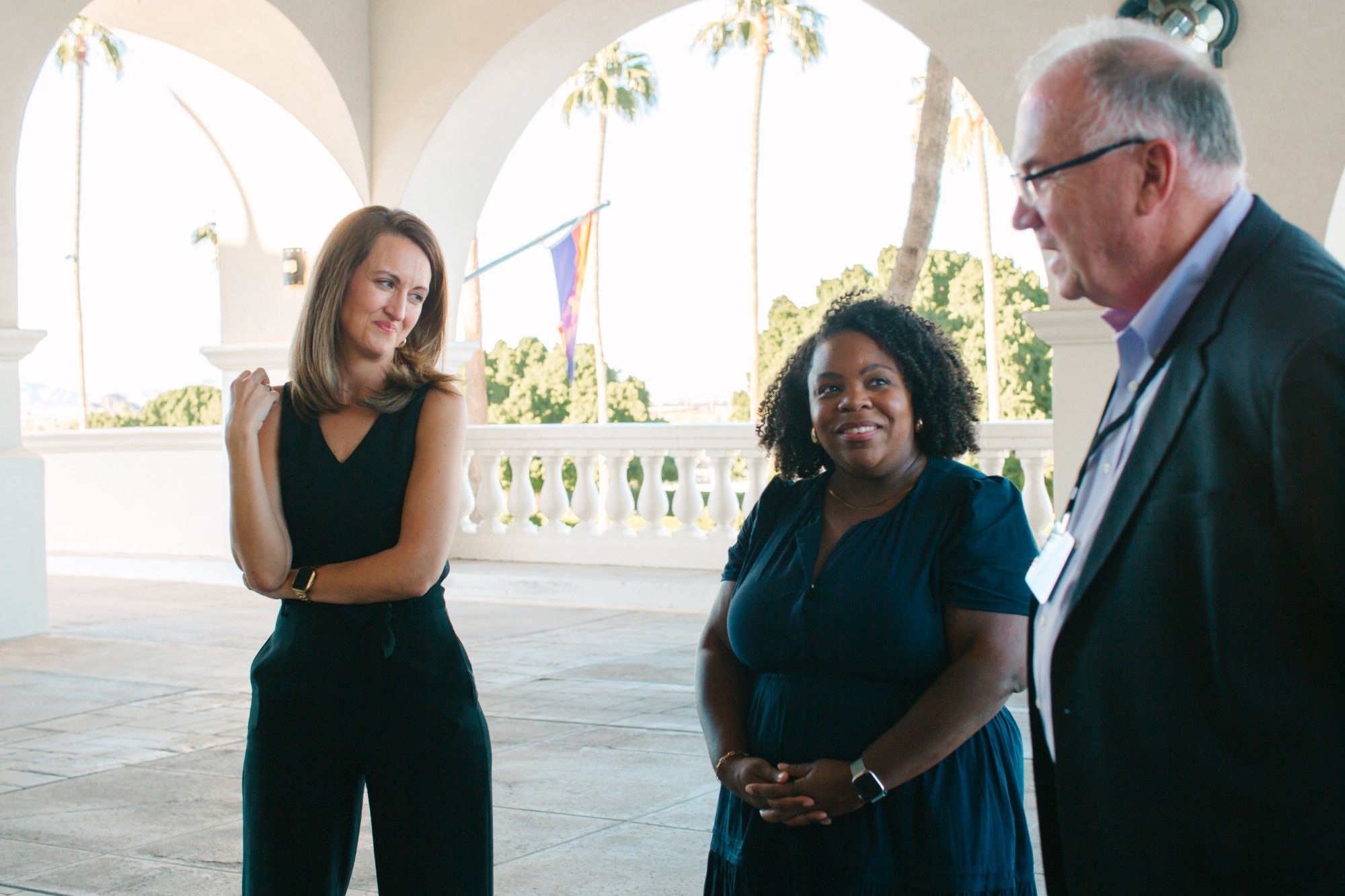 Three educators talk on a sunlit arched terrace with palm trees beyond, two women smiling as a man in a dark blazer and lanyard speaks