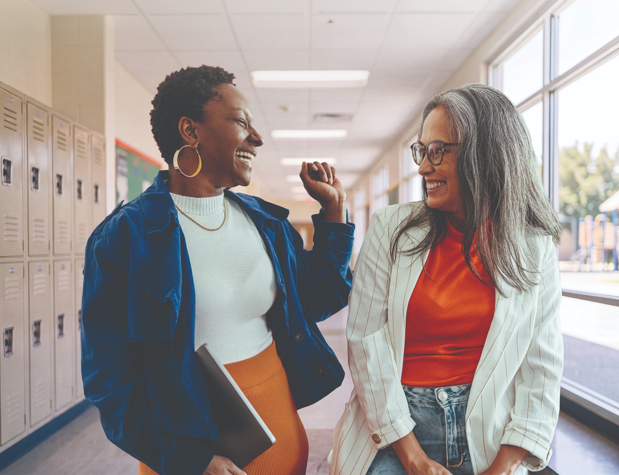 Two educators laughing together in a sunlit school hallway, one in a cobalt jacket holding a laptop, the other in a striped blazer and glasses, lockers on one side and windows on the other