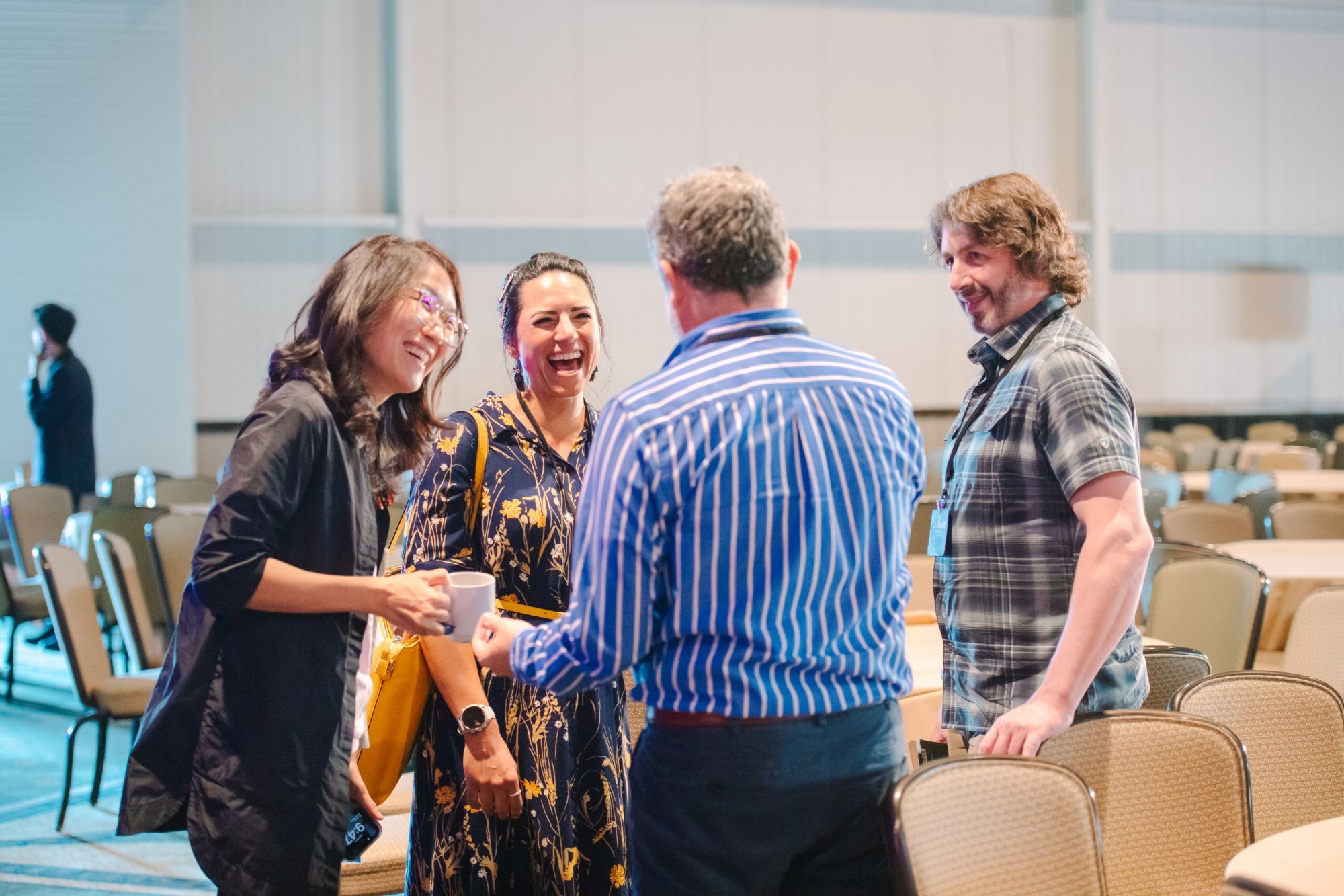 Four educators with lanyards share a laugh between sessions in a conference hall, one holding a coffee mug, rows of empty chairs and round tables behind them