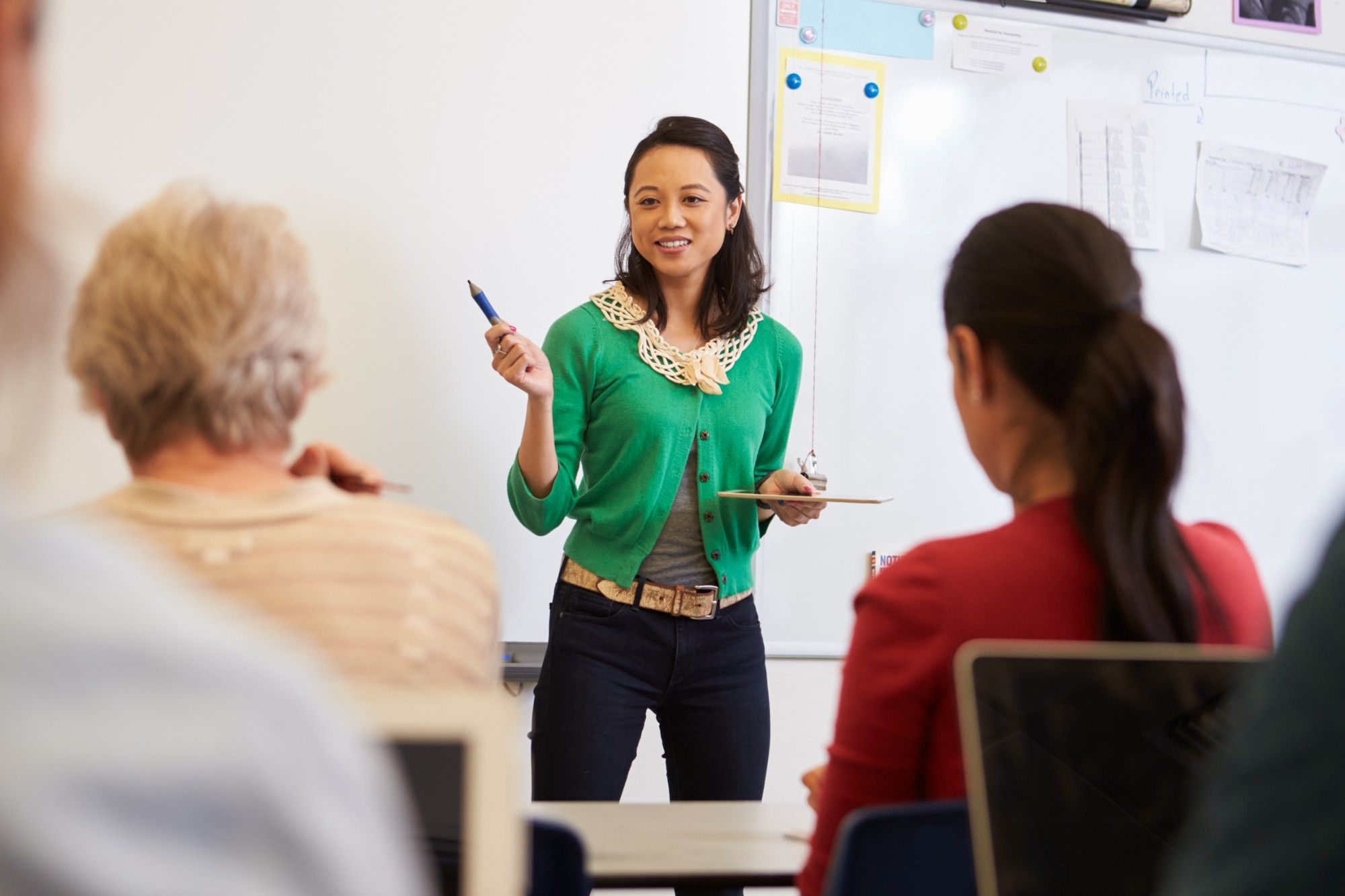 A smiling facilitator in a green cardigan, pen and tablet in hand, leads a session at a whiteboard while educators at laptops listen, seen from behind
