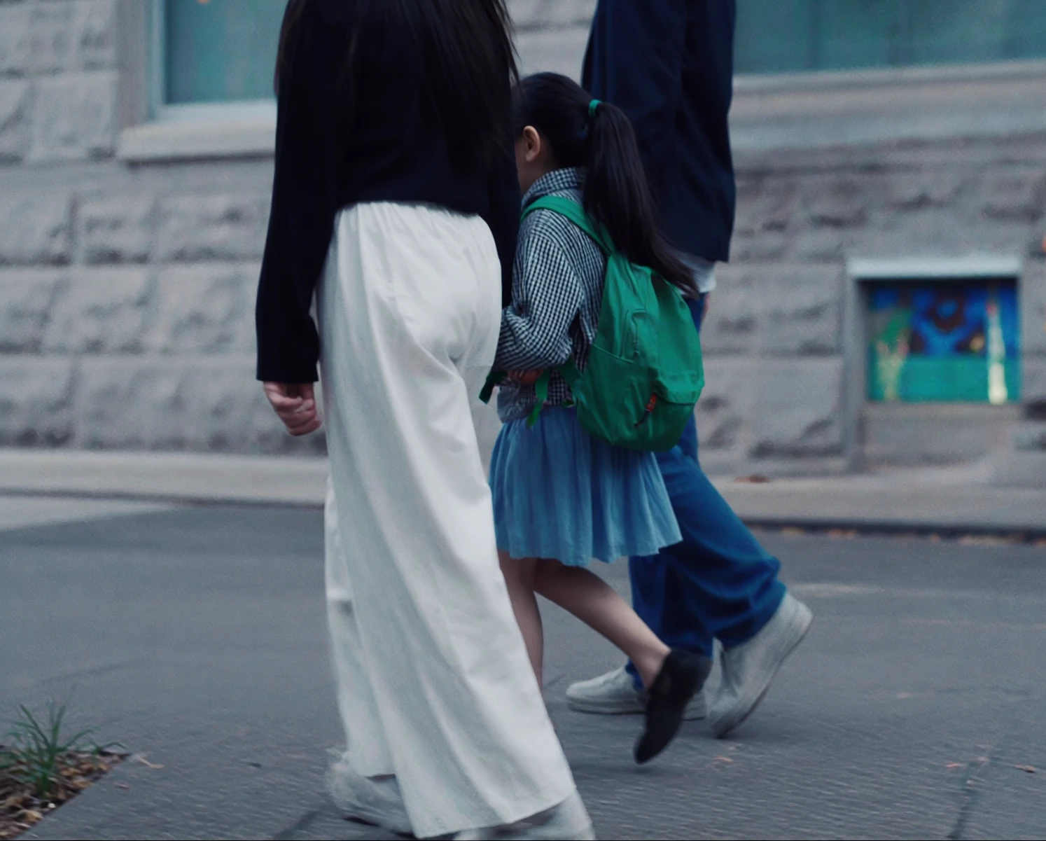 A girl in a gingham shirt and blue skirt, a green backpack on her shoulders, walks to school between her mother and father past a stone wall