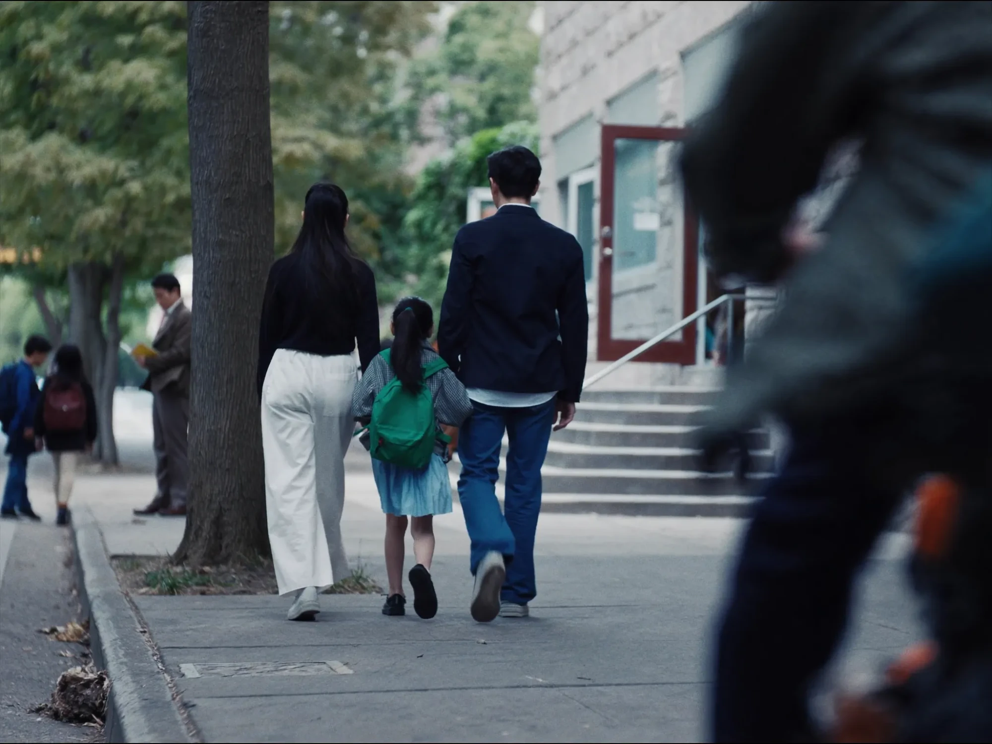 A mother and father walk their young daughter, a green backpack on her shoulders, along a tree-lined sidewalk toward the steps and red front doors of a stone school building