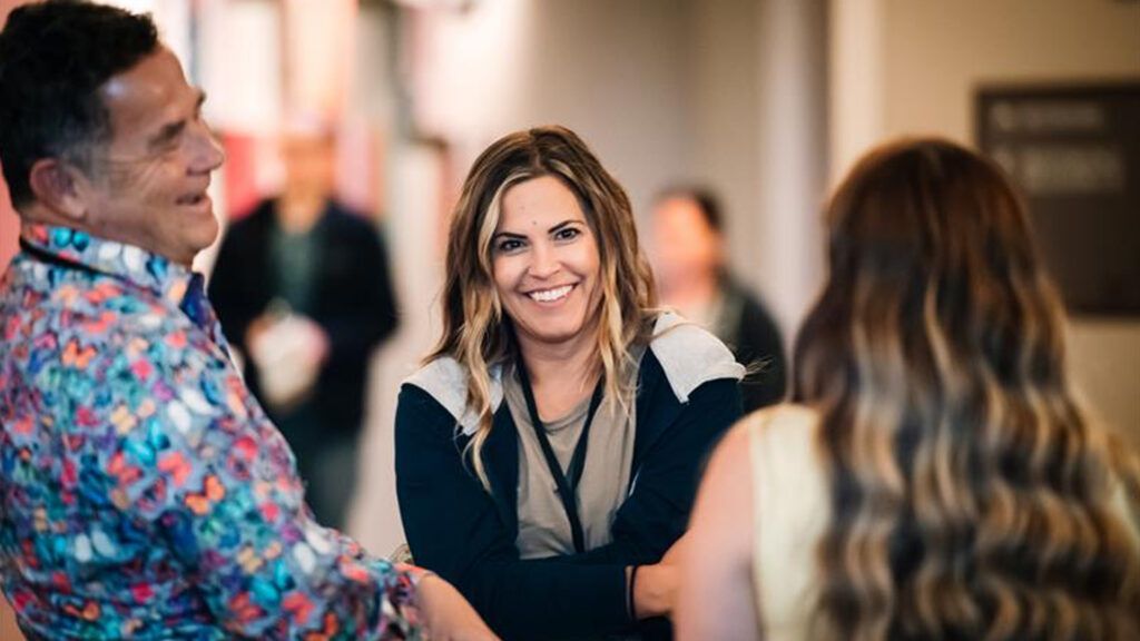 Two conference attendees greet each other with smiles in a hallway, one in a bright butterfly-print shirt