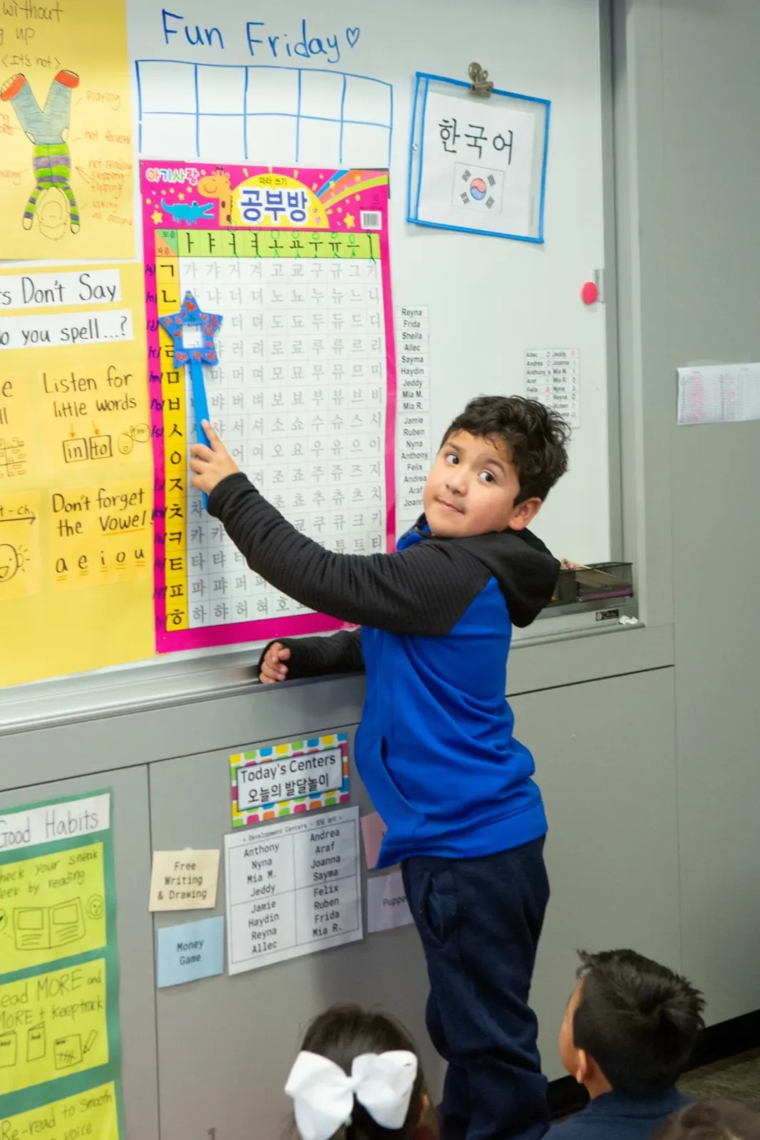 A young student points to a word on a classroom chart labeled “Fun Friday,” beside signs in Korean