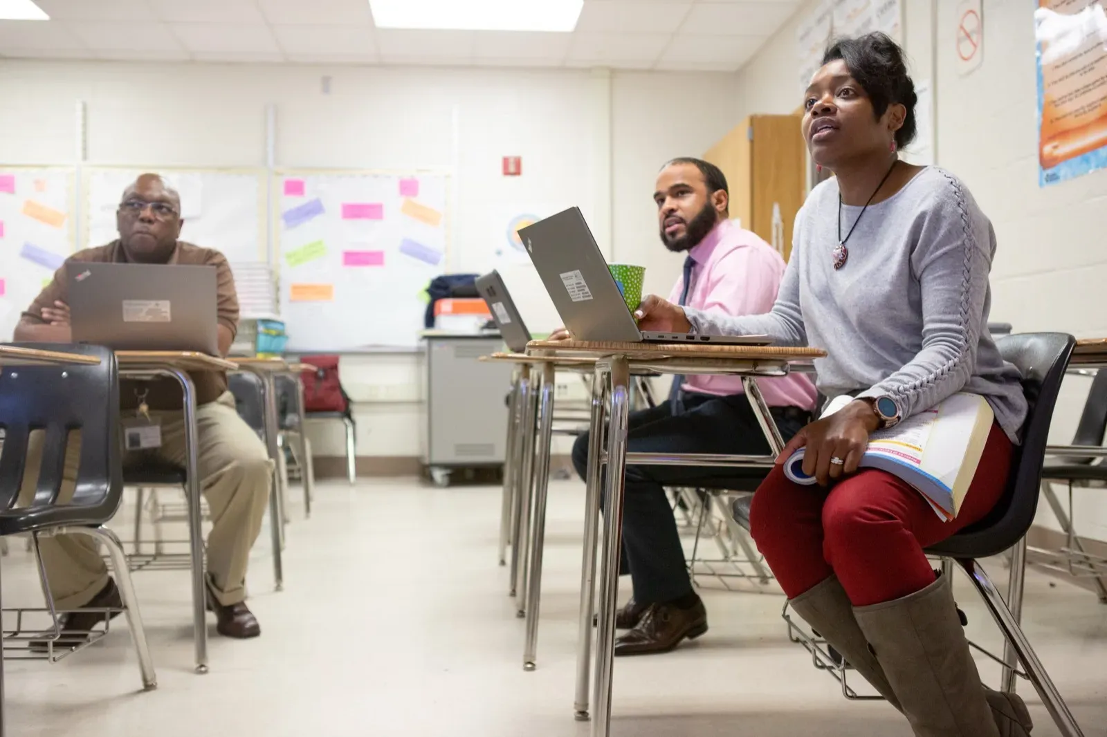 Educators seated at classroom desks with laptops look toward the front of the room during a session
