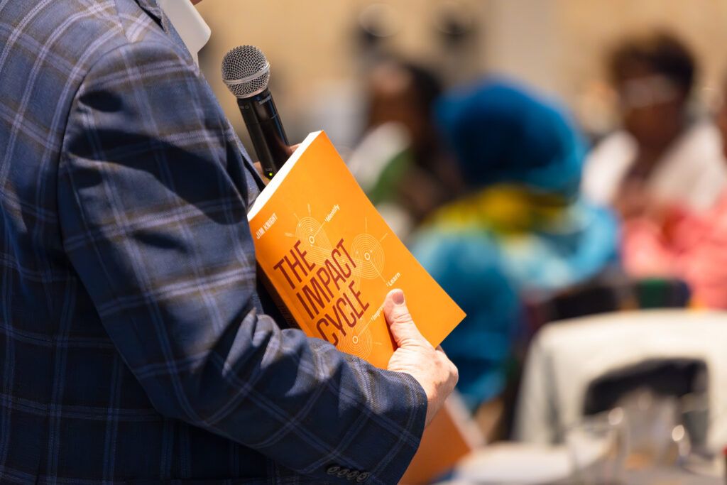 Jim Knight, in a plaid blazer, holds a microphone and a copy of his book The Impact Cycle as he speaks, the audience blurred behind