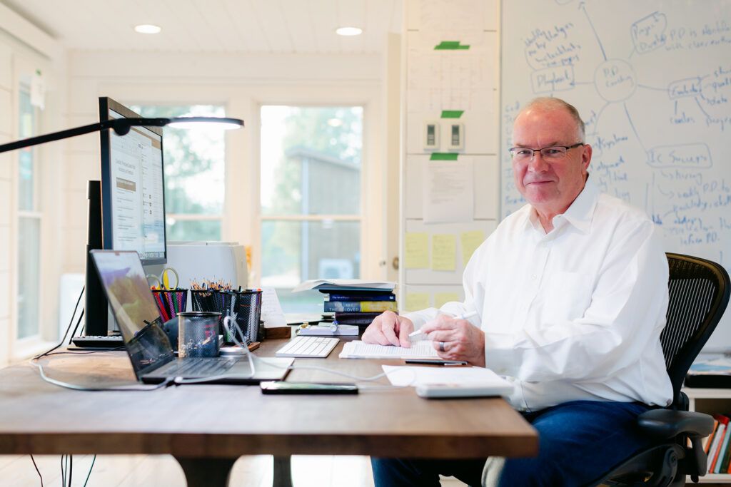 Jim Knight at his desk in a white shirt and glasses, pen in hand over a notebook, a whiteboard of handwritten coaching notes behind him
