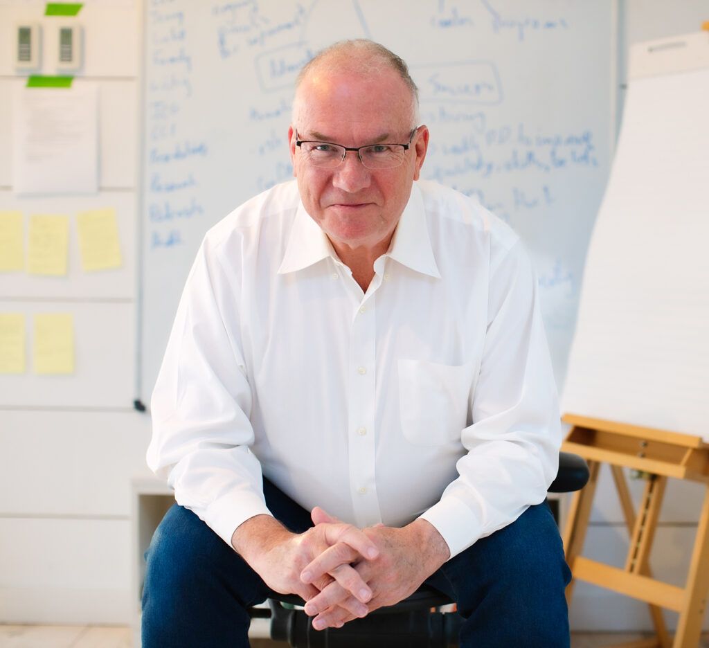 Jim Knight, in a white shirt and glasses, sitting forward with his hands clasped in front of a whiteboard covered in handwritten notes
