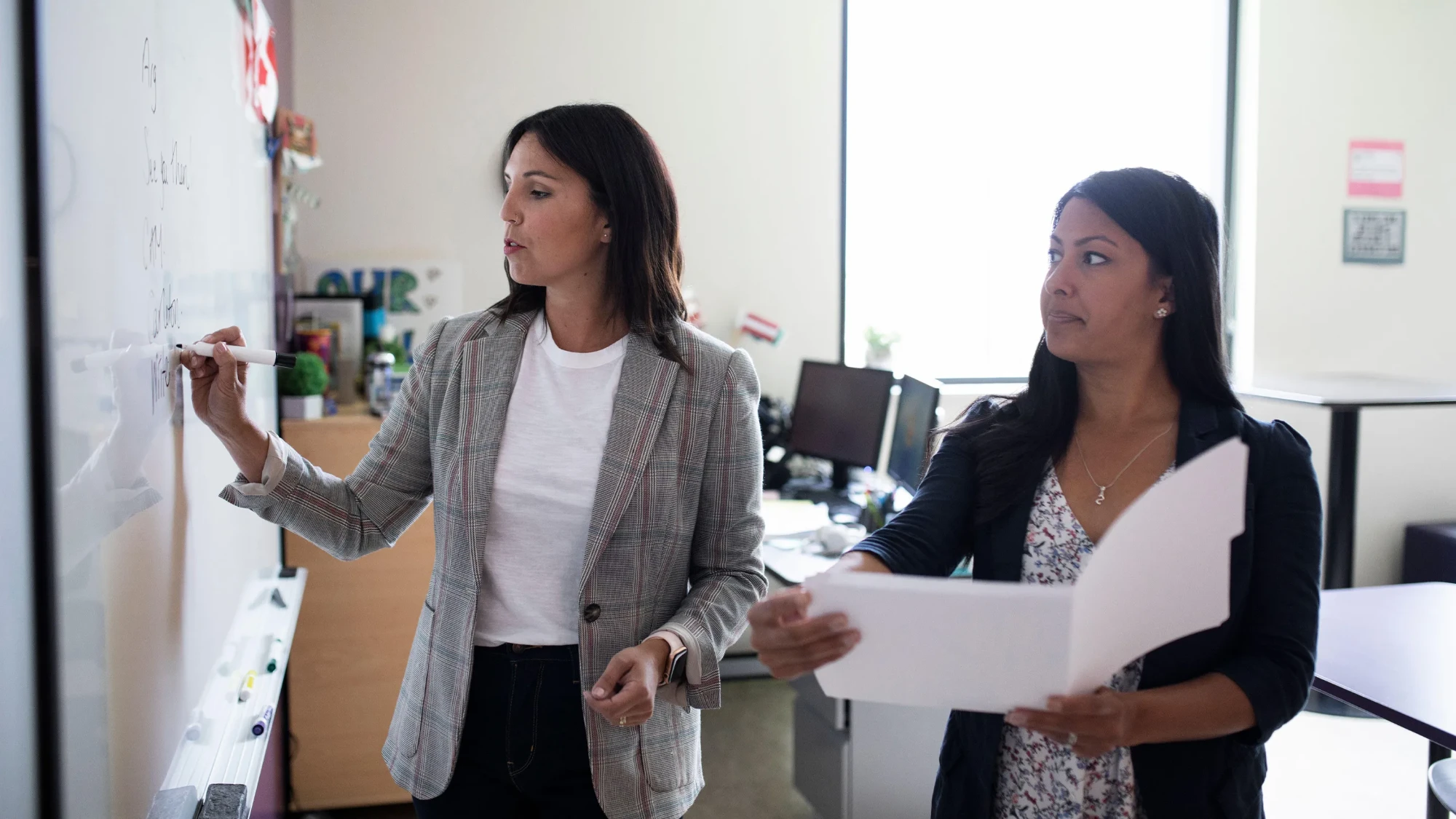 An educator in a plaid blazer writes on a whiteboard while a colleague holding an open folder of papers stands beside her and reads along, desks and monitors in the office behind them