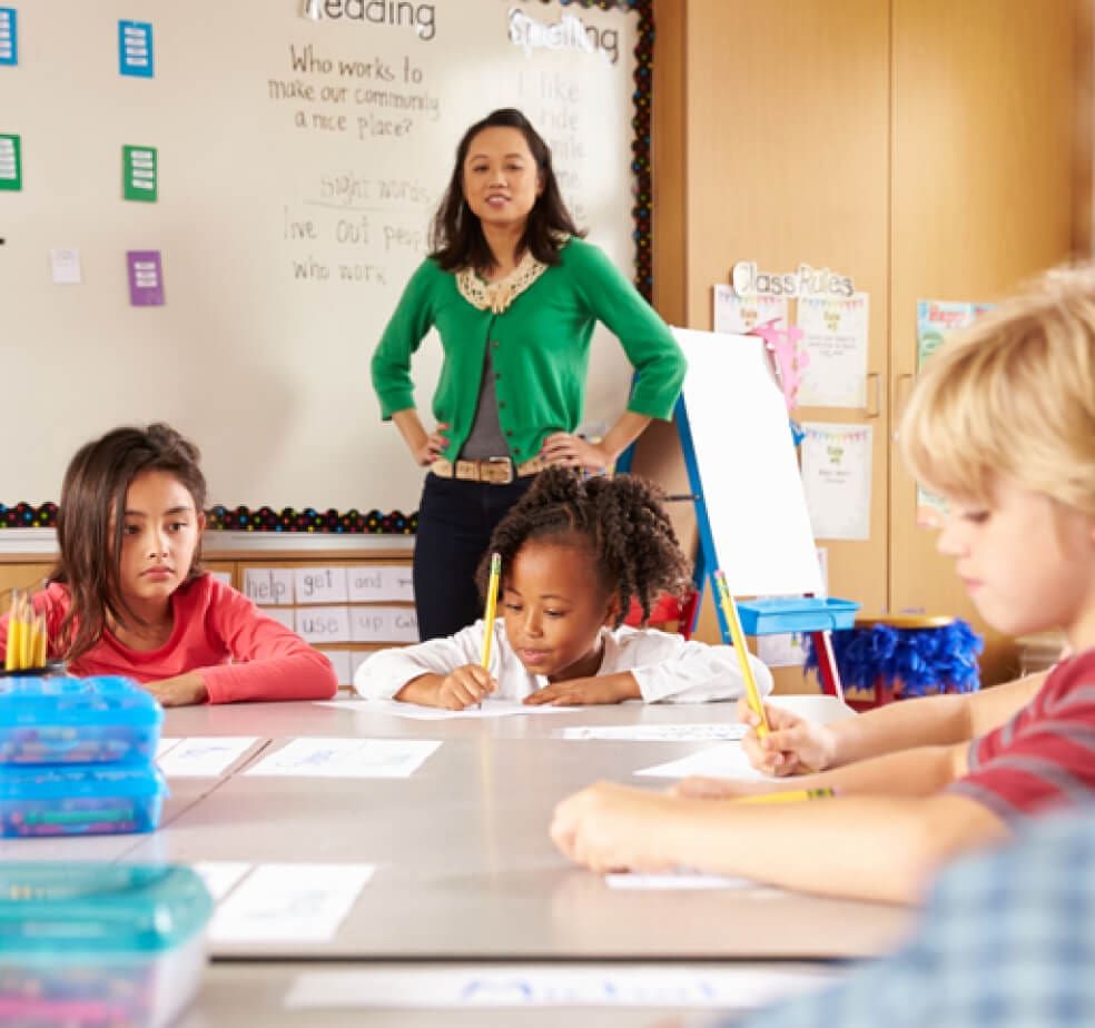 An elementary teacher in a green cardigan stands at the front of a classroom while students at a long table write on worksheets, a reading and sight-words chart on the whiteboard behind