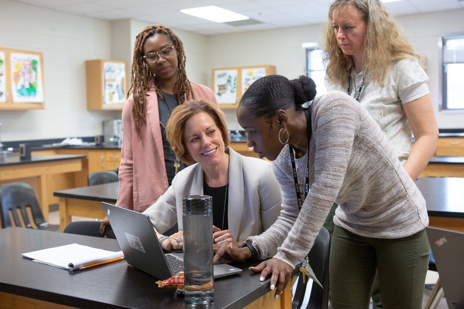 Four educators in a science classroom gather around a laptop, one seated and smiling while a colleague in a lanyard leans in to point at the screen and two others look on