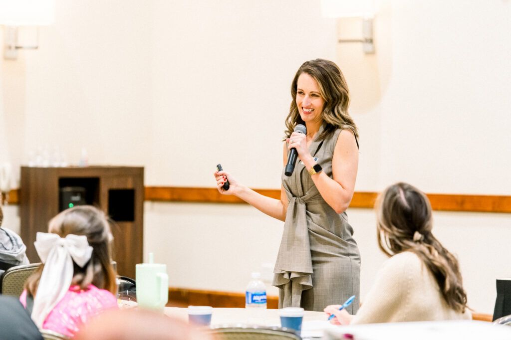 A presenter in a gray dress smiles as she speaks into a microphone with a clicker in her other hand, attendees at tables in front of her