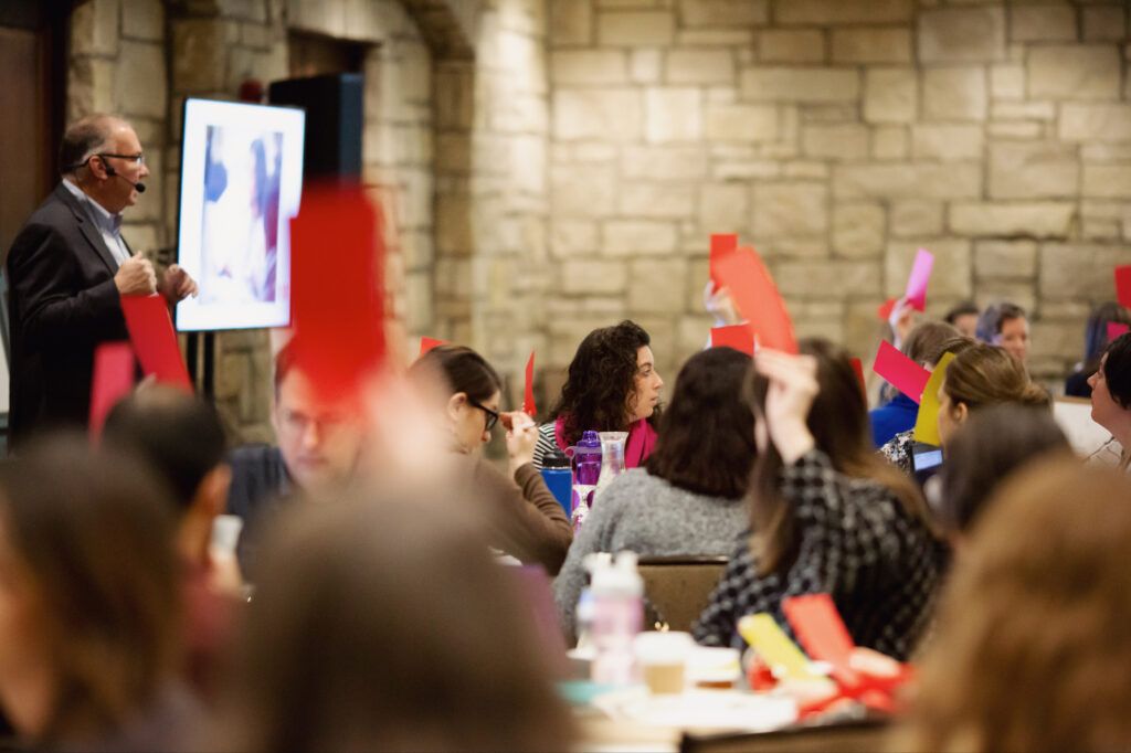 Attendees at round tables hold up red, pink and yellow response cards while a presenter with a headset microphone speaks beside a screen