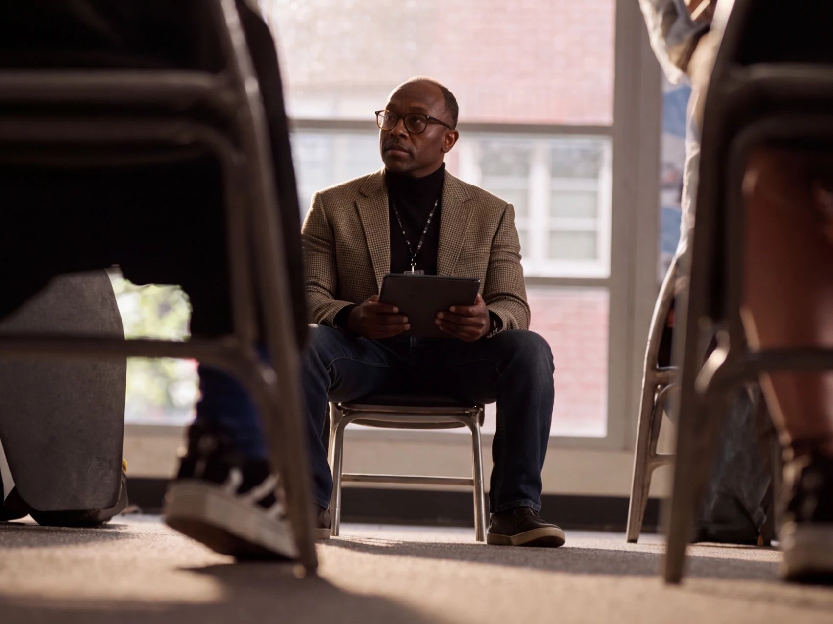 A school leader in a tweed jacket and glasses sits with a tablet in a circle of chairs, listening in front of a sunlit window