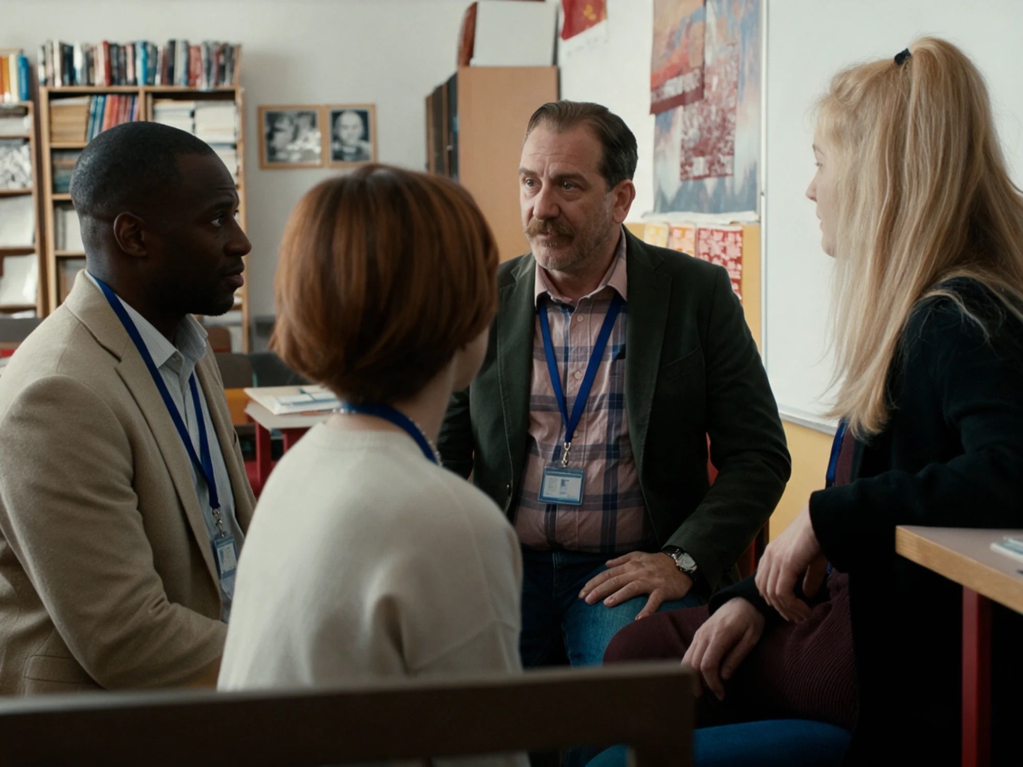 Four educators wearing lanyards talk in a circle of chairs in a classroom, a man in a dark blazer leaning in to listen as a colleague speaks, bookshelves and a whiteboard behind them