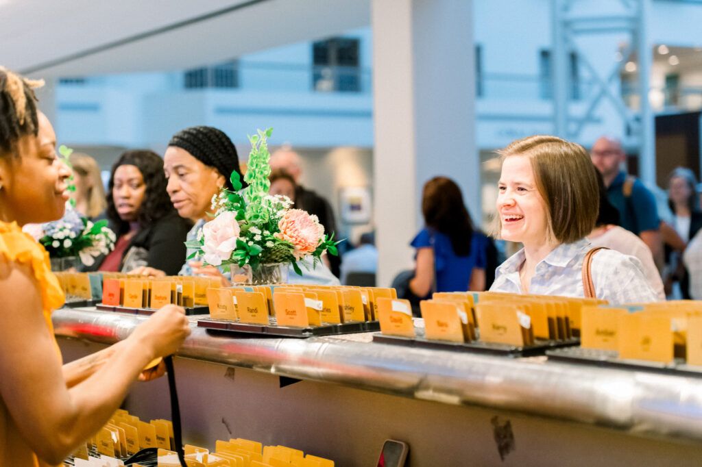 Attendees laugh and talk at a conference registration desk lined with rows of printed name badges and a vase of flowers