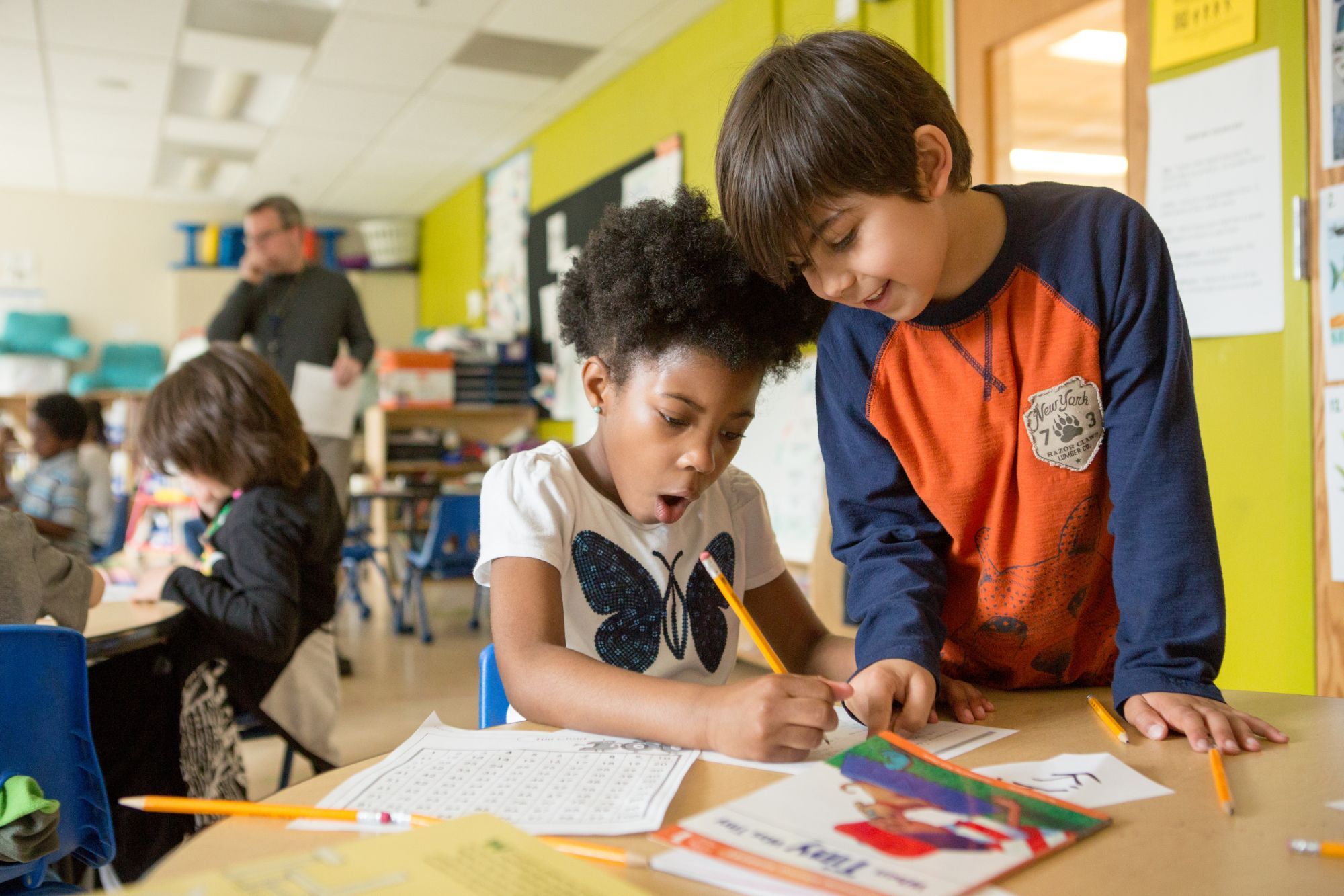 Two young students at a classroom table, a girl writing on a worksheet with a pencil as a boy leans over her shoulder pointing to the page, a teacher and classmates working behind them
