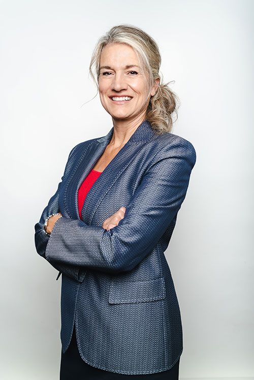 Shannon K. Buerk, in a navy blazer over a red top, smiling with her arms folded against a light gray backdrop