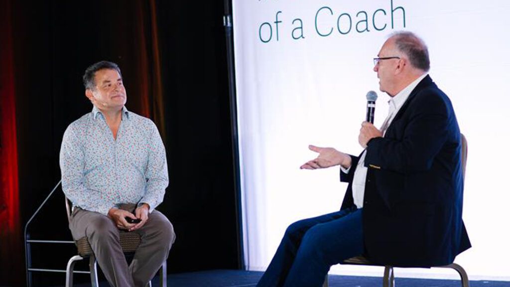 Two speakers seated on a conference stage, one holding a microphone and gesturing as the other listens