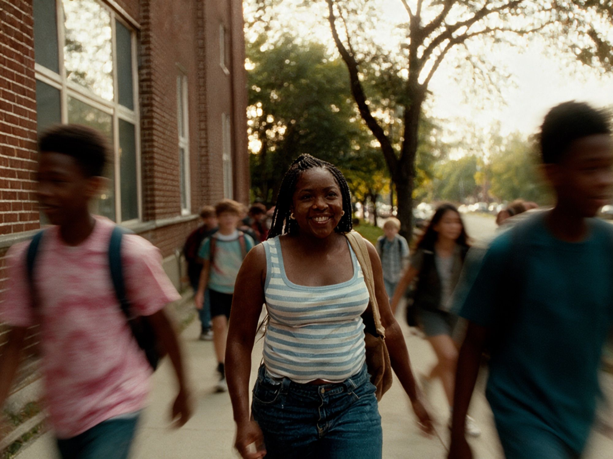 A smiling student with braids, in a striped tank top with a canvas bag on her shoulder, walks along a sidewalk beside a brick school building as classmates hurry past