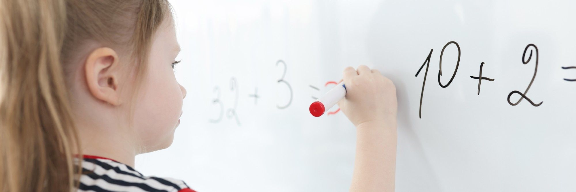 Seen from behind her shoulder, a young student in a striped shirt writes an answer in red marker beside 32 + 3 on a whiteboard, 10 + 2 written larger beside it