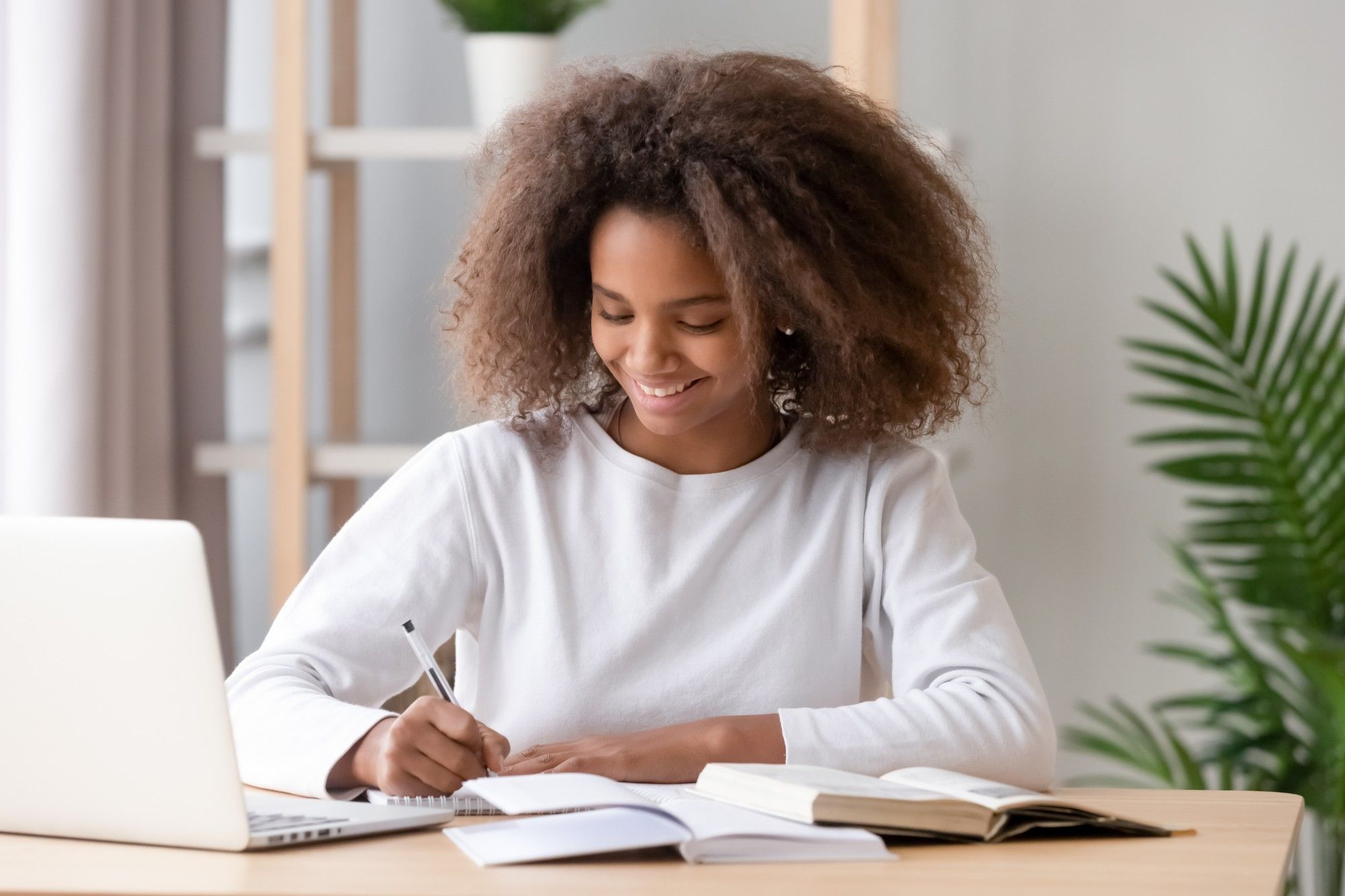 A smiling student with curly hair, in a white sweater, writes in a notebook at a desk beside an open laptop and an open book, a palm plant behind her