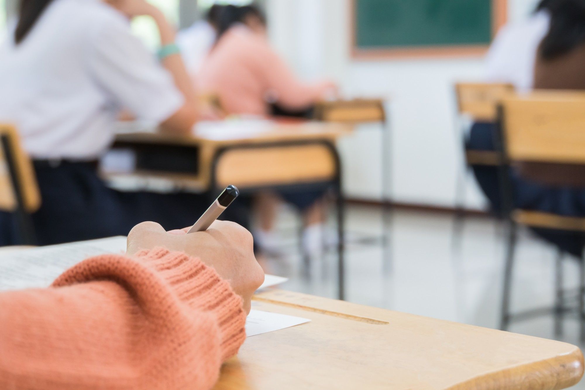 Close on a student’s hand in a coral sweater writing with a pencil at a wooden desk, classmates working at desks out of focus ahead
