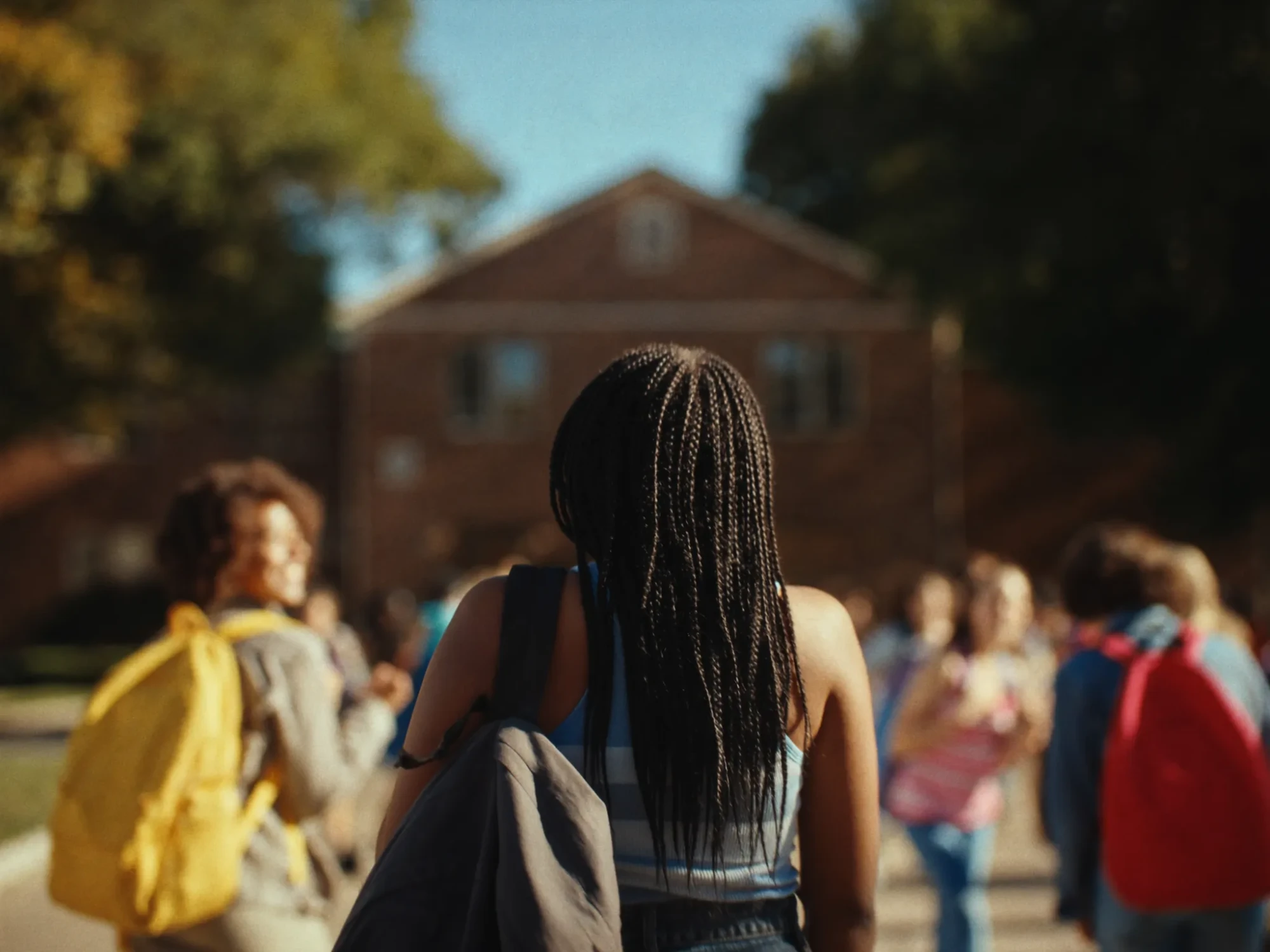 Seen from behind, a student with long braids and a gray backpack walks toward a brick school building on a sunny morning, classmates with yellow and red backpacks around her
