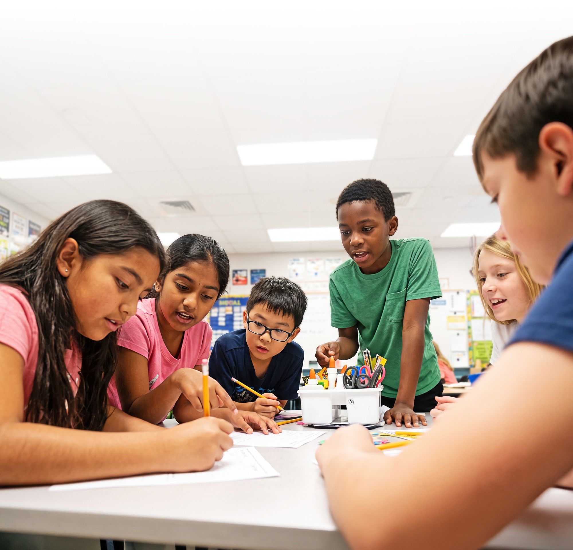 Five students lean over a classroom table working through a worksheet together, one pointing to the page while another writes
