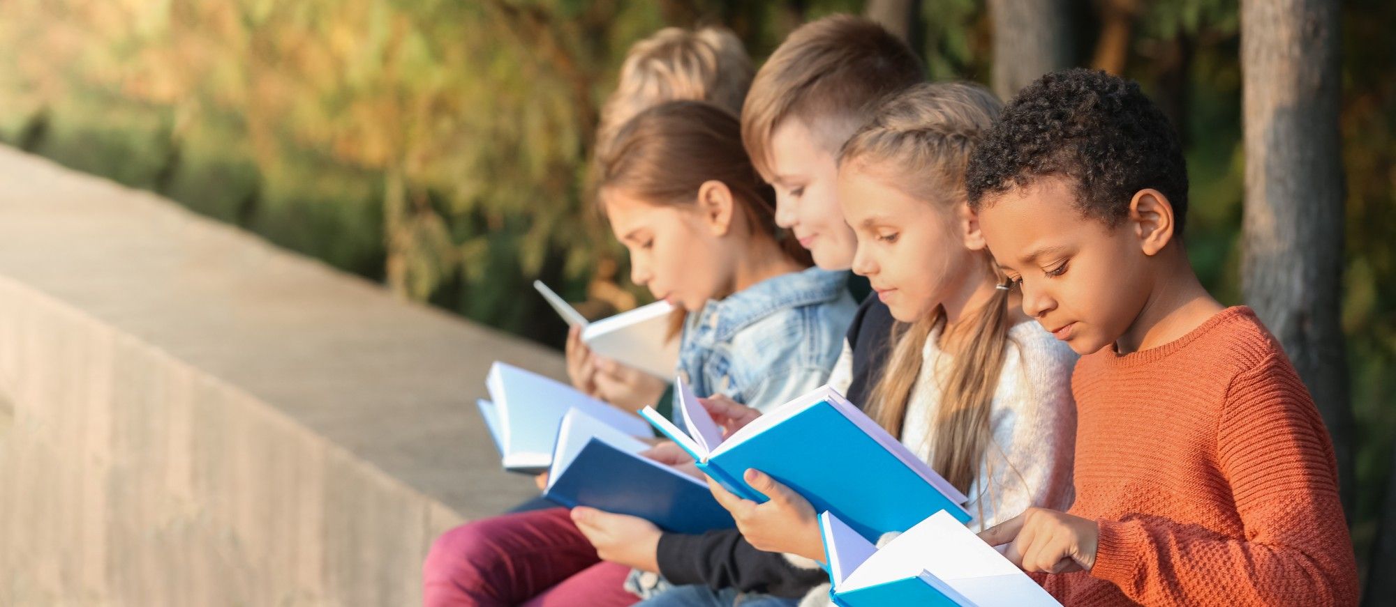 Five young students sit side by side on a low stone wall outdoors in autumn light, each reading an open blue book, a boy in an orange sweater pointing to a line on his page