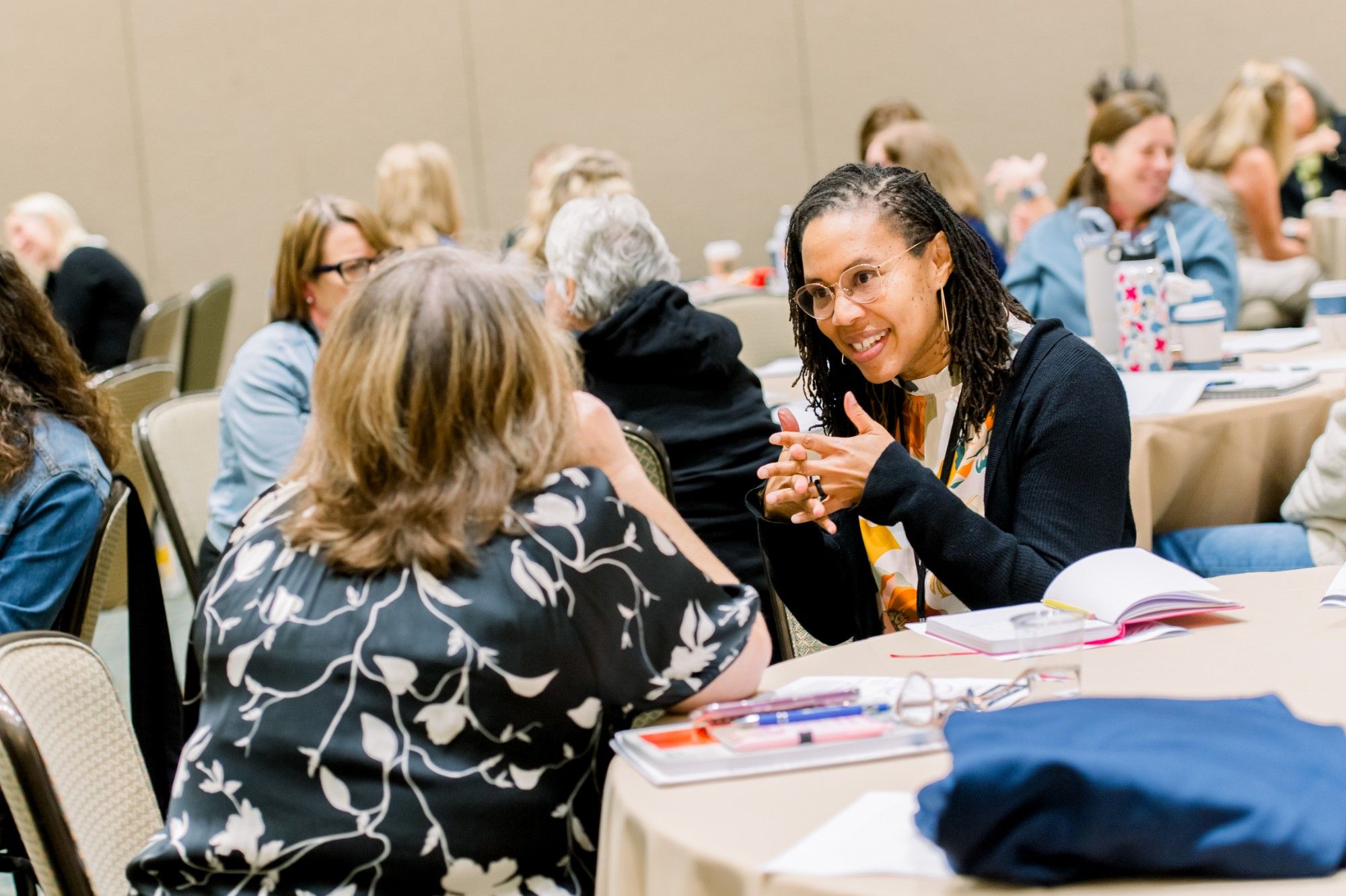 At a round table in a busy conference hall, a smiling educator in glasses and a navy cardigan gestures while talking with a colleague seen from behind, notebooks and pens on the table and other tables of educators out of focus behind them