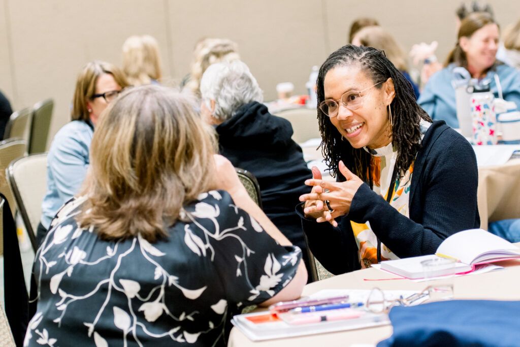 An educator in glasses, her locs pulled back, talks with her hands to a colleague across a conference table, attendees at round tables behind them