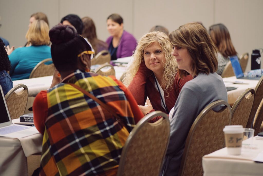 Three educators lean in to talk at a conference table, others working at round tables behind them