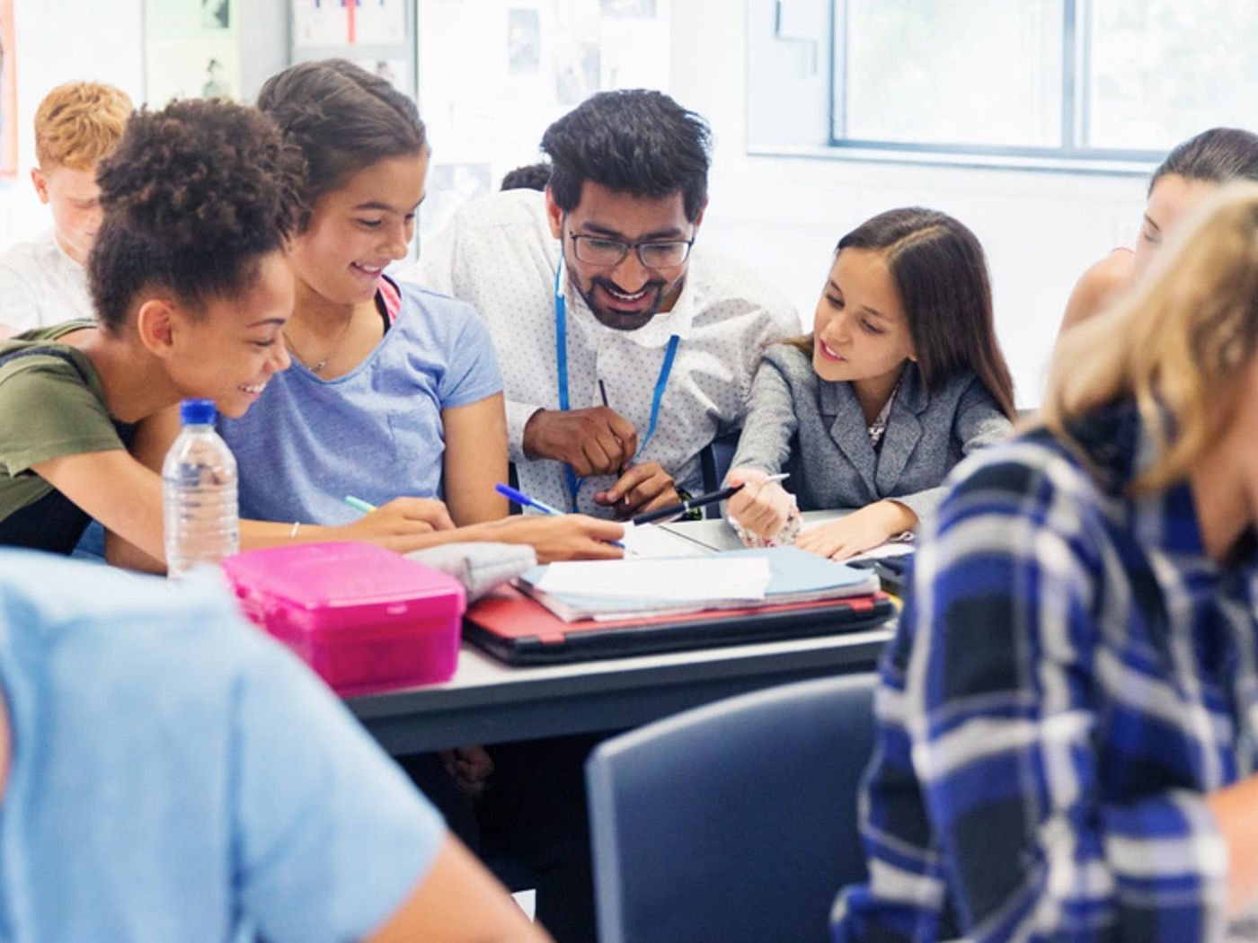 A smiling teacher in glasses and a lanyard leans in over a table of open notebooks with three students, pointing out something on the page as classmates work nearby