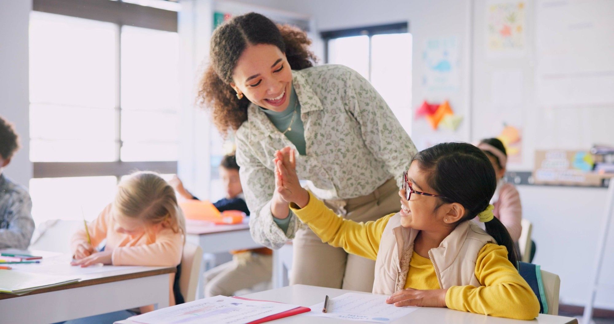 A smiling teacher in a floral blouse leans down to high-five a laughing student in glasses and a yellow sweater at her desk, classmates working around them