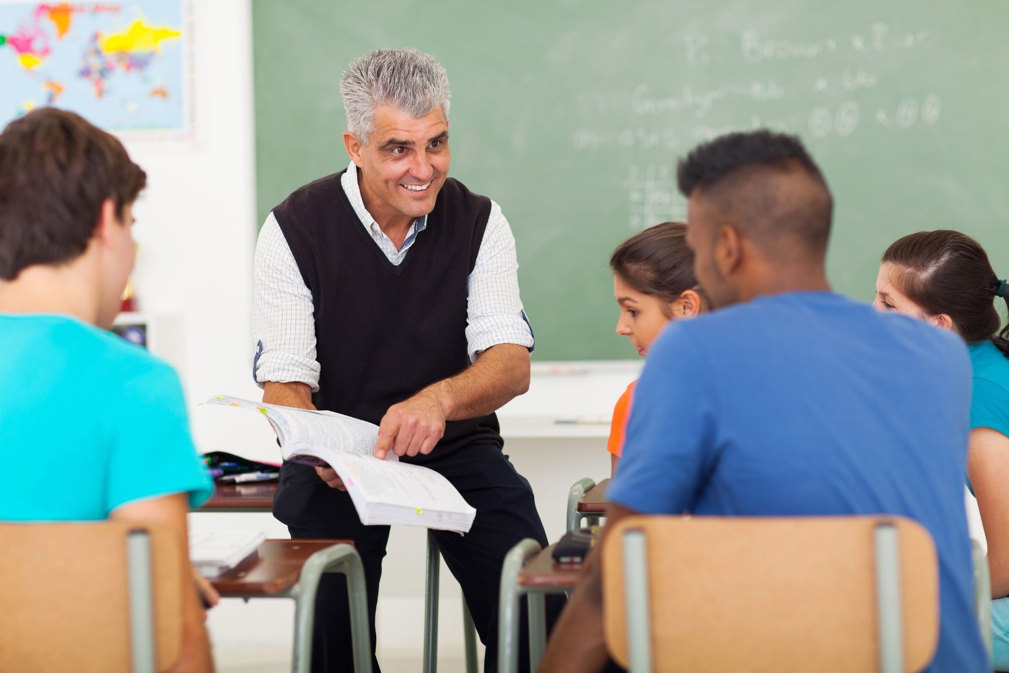 A smiling teacher with gray hair, in a black sweater vest, leans in with an open textbook to talk with a small group of high school students at their desks, a chalkboard behind him