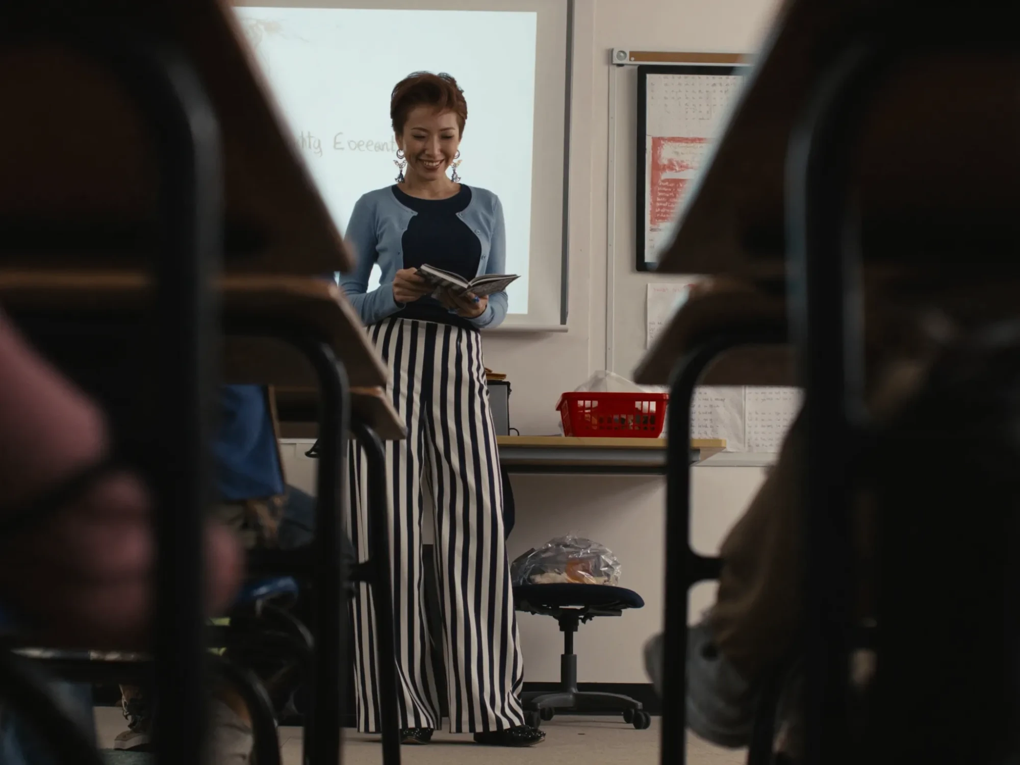 Seen between the desks from a student’s seat, a smiling teacher in a blue cardigan and striped trousers reads from a book at the front of the classroom