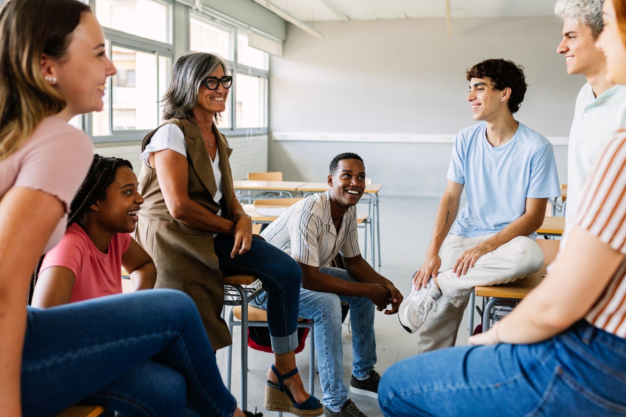 A smiling teacher with gray hair and glasses sits on a desk in a circle of laughing high school students, talking together in a bright classroom