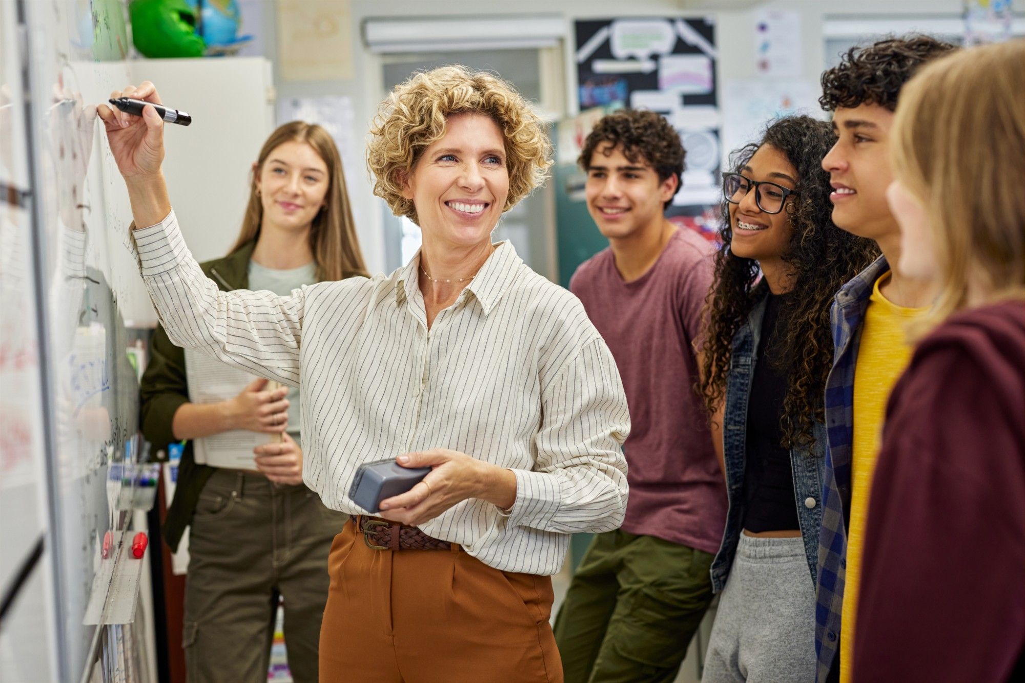A smiling teacher with curly hair, in a striped shirt, writes on a whiteboard, marker and eraser in hand, as a group of smiling high school students looks on
