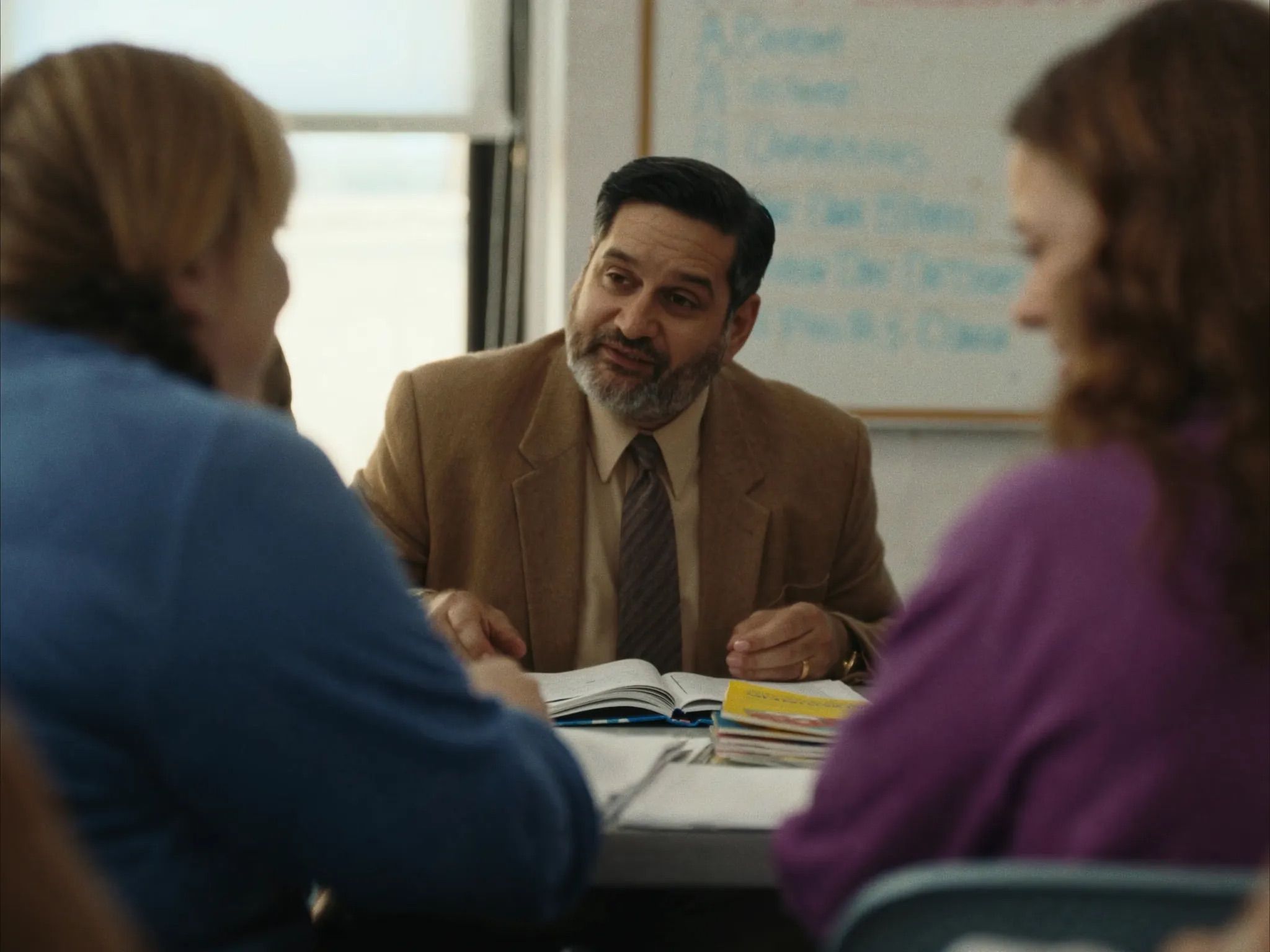 A bearded educator in a tan jacket and tie talks with two teachers across a table of open curriculum books, a whiteboard behind him
