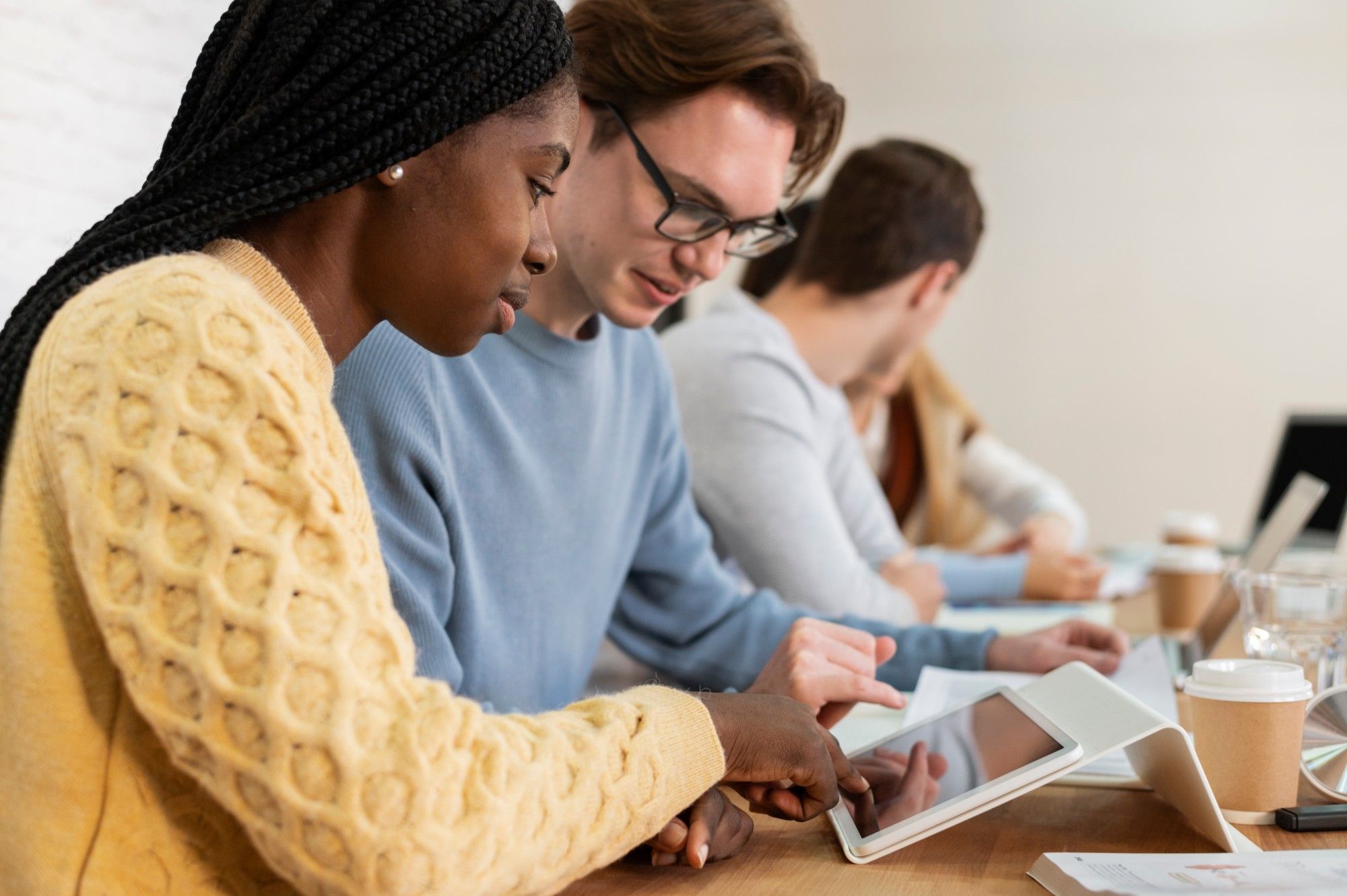 Two educators, one in a yellow cable-knit sweater and one in glasses and a blue sweater, review a tablet together at a long meeting table, colleagues with laptops and coffee beside them