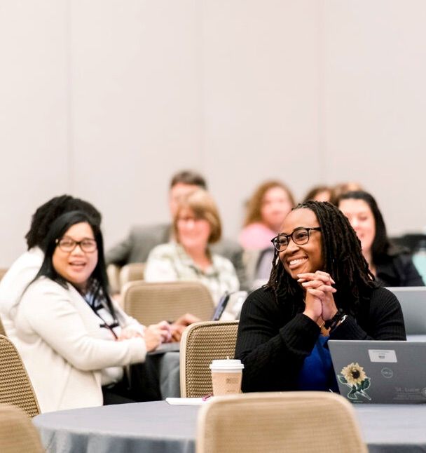 Educators seated at round tables in a conference session smile toward the front of the room, one in the foreground with hands clasped beside a laptop