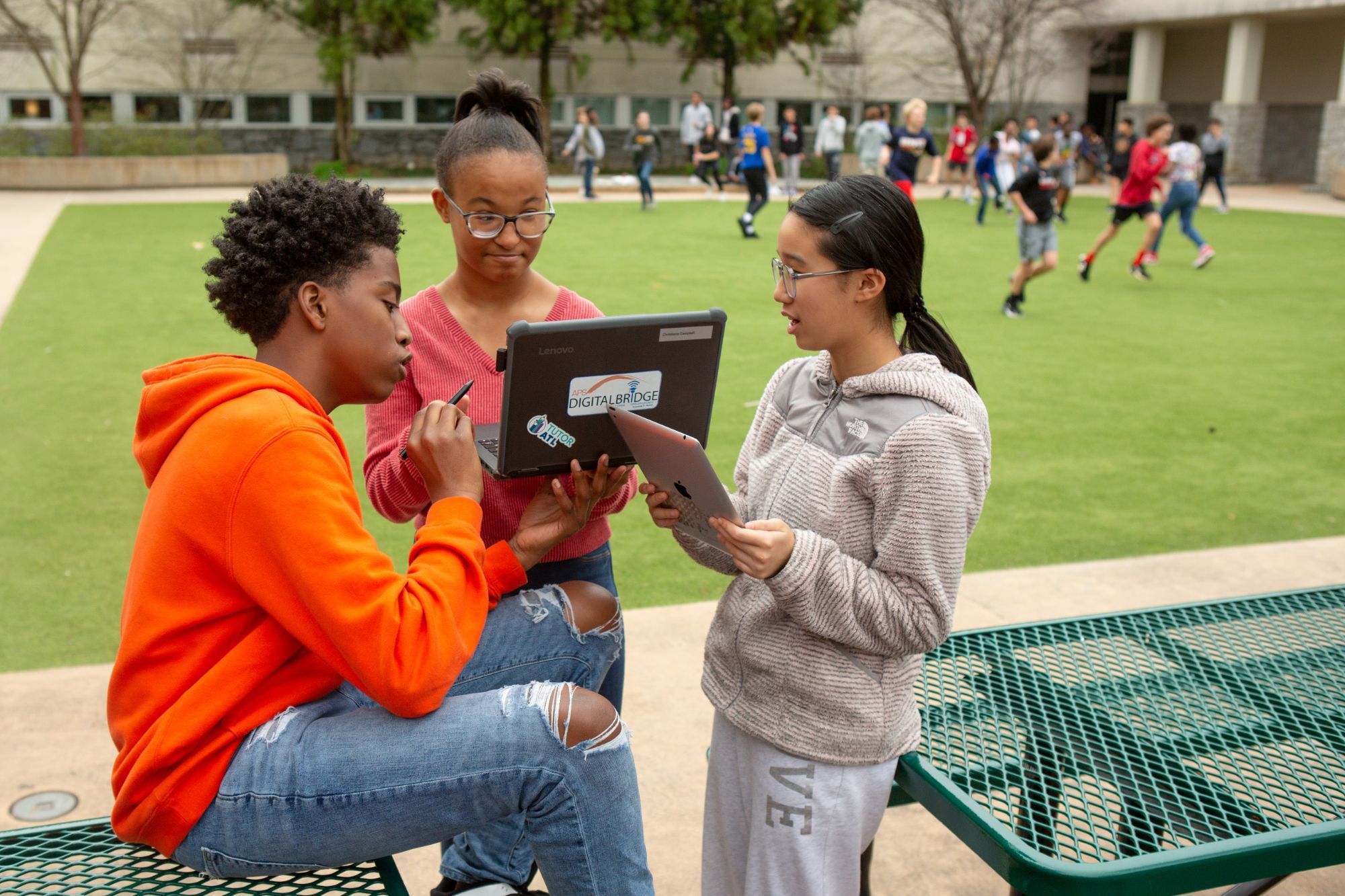 Three middle school students talk through their work at a courtyard picnic table, one seated with a stylus, one holding up a laptop and one with a tablet, classmates running across the lawn behind them